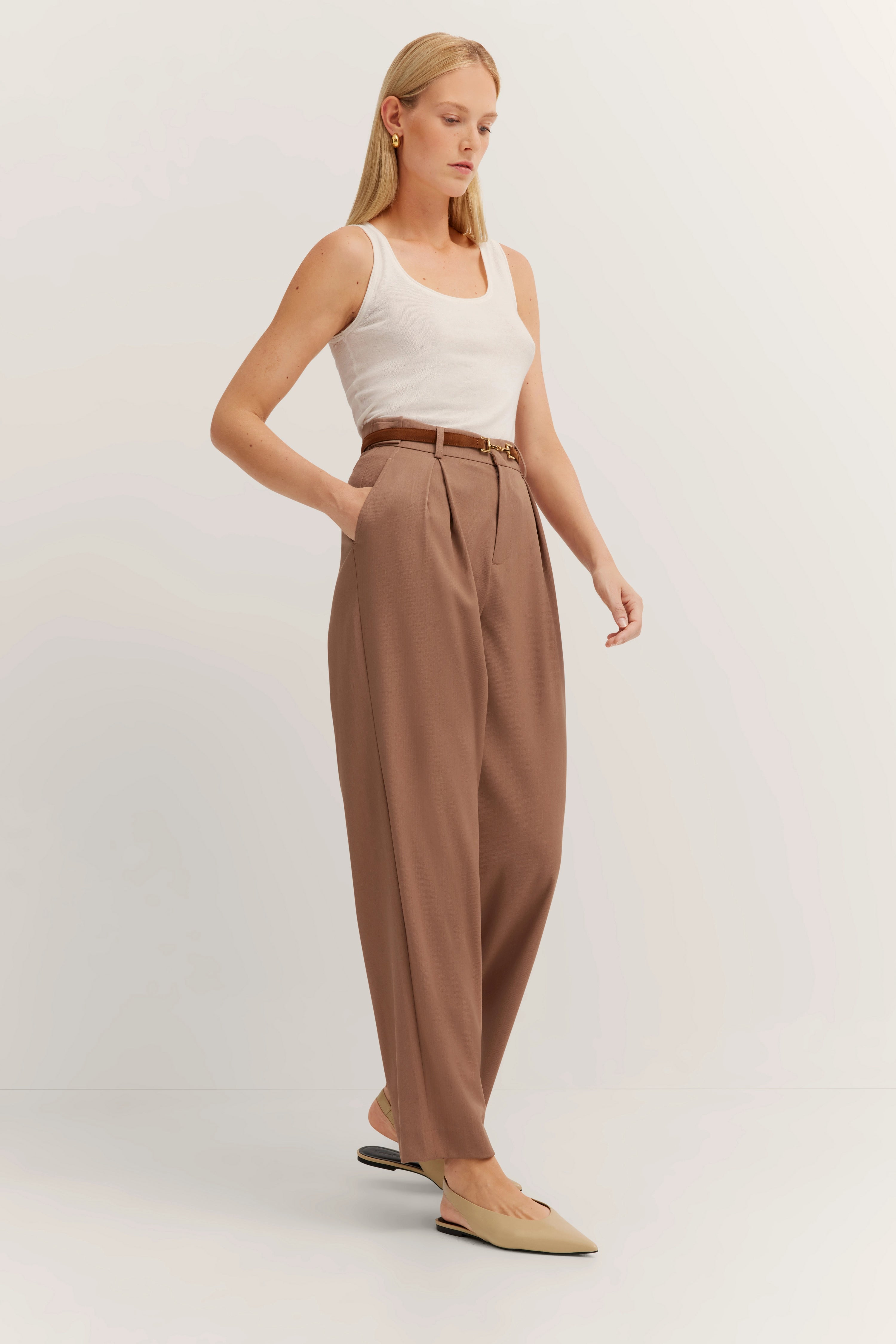Beige model met bruine broek, ontspannen pose, wit topje en sandalen, straalt bugatti's stijlvolle eenvoud uit.