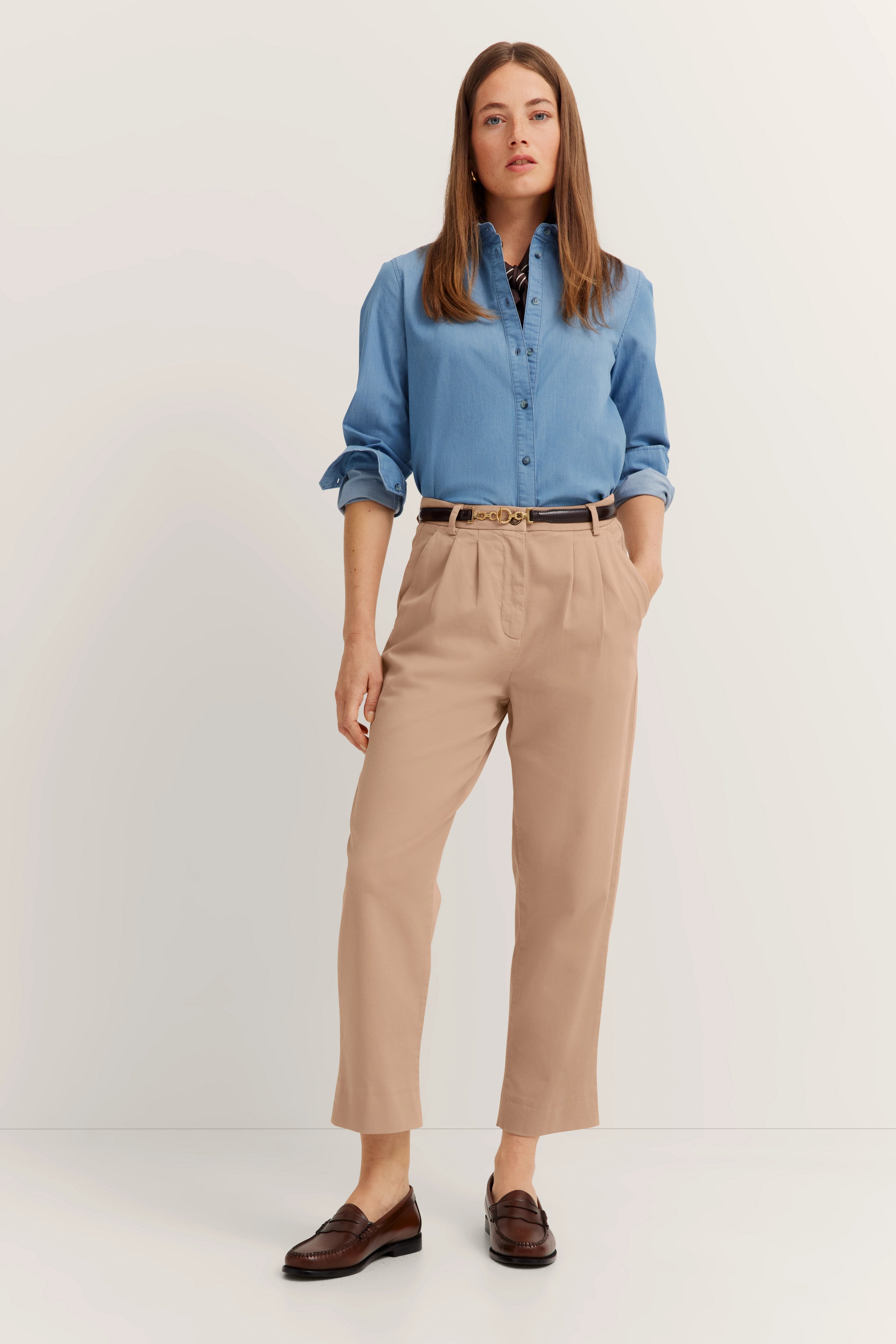 Model draagt blauw overhemd, beige broek, bruine loafers, handen in broekzakken. Bugatti-stijl en elegantie.