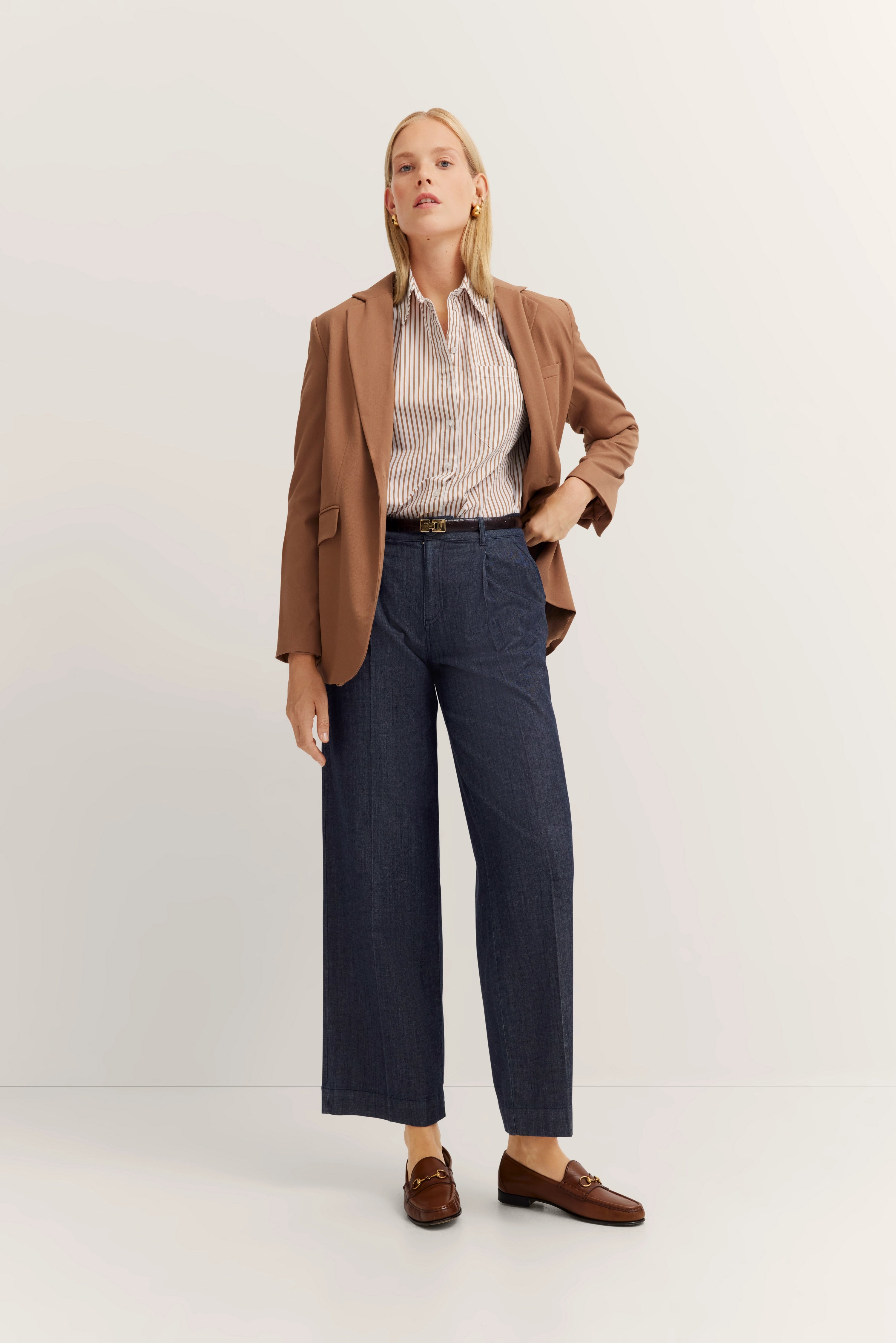Elegant modelbeeld: vrouw in bruine blazer, gestreepte blouse, blauwe broek, bruine loafers, zelfverzekerde pose.