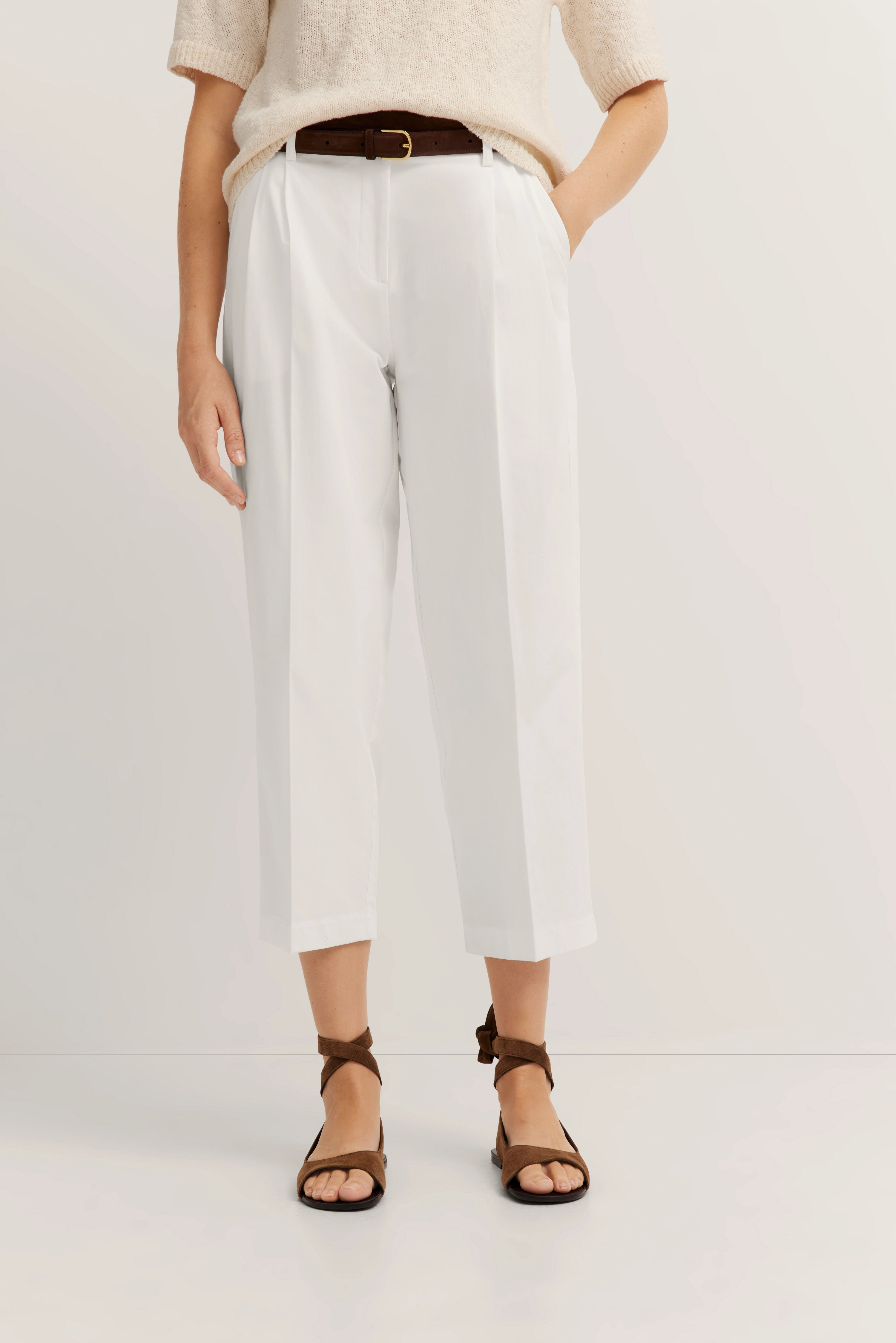 Beige trui, witte broek, bruine riem en sandalen, casual modelbeeld, ontspannen pose.