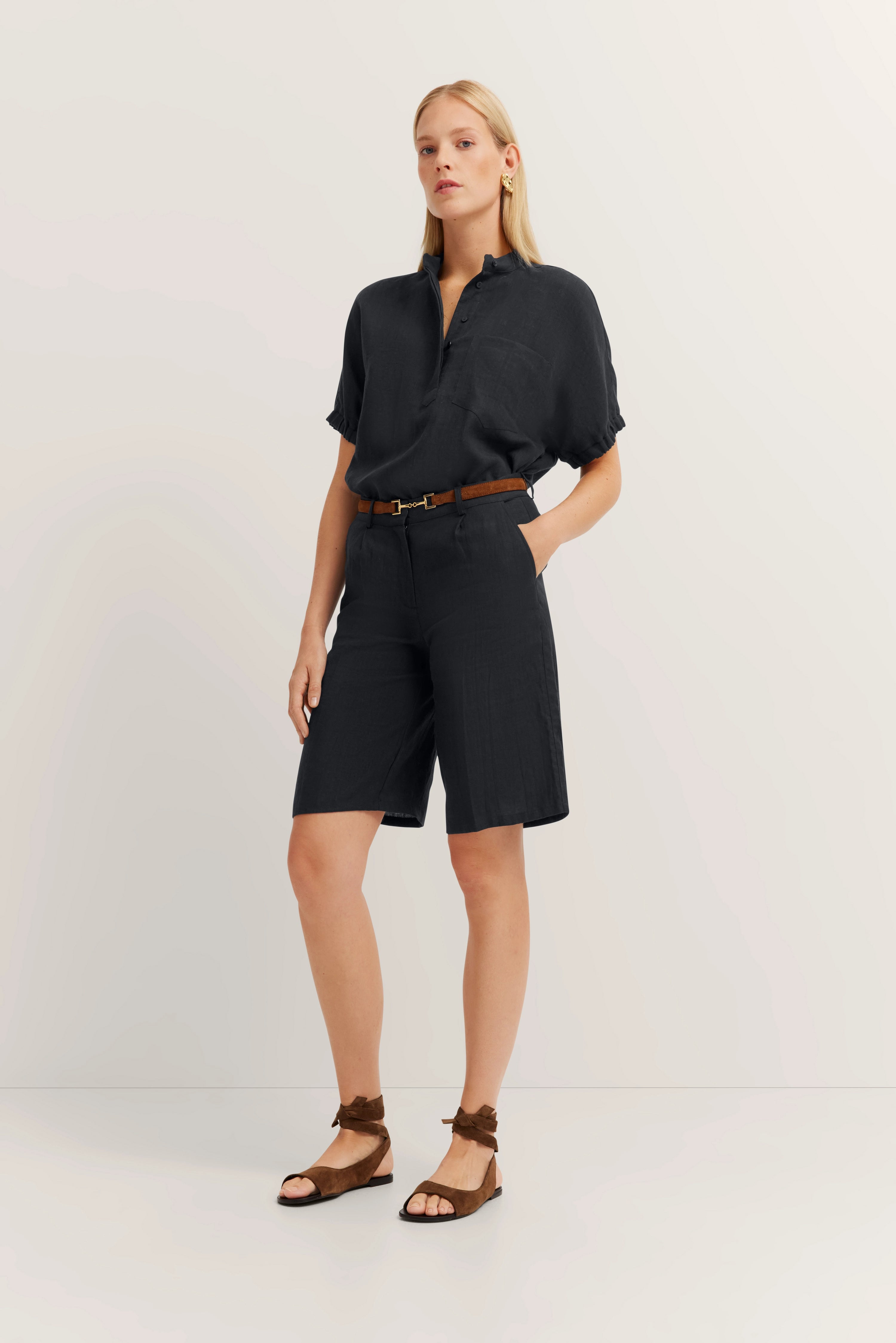Model in zwarte outfit met blouse en bermudashorts, bruine riem, ontspannen pose.