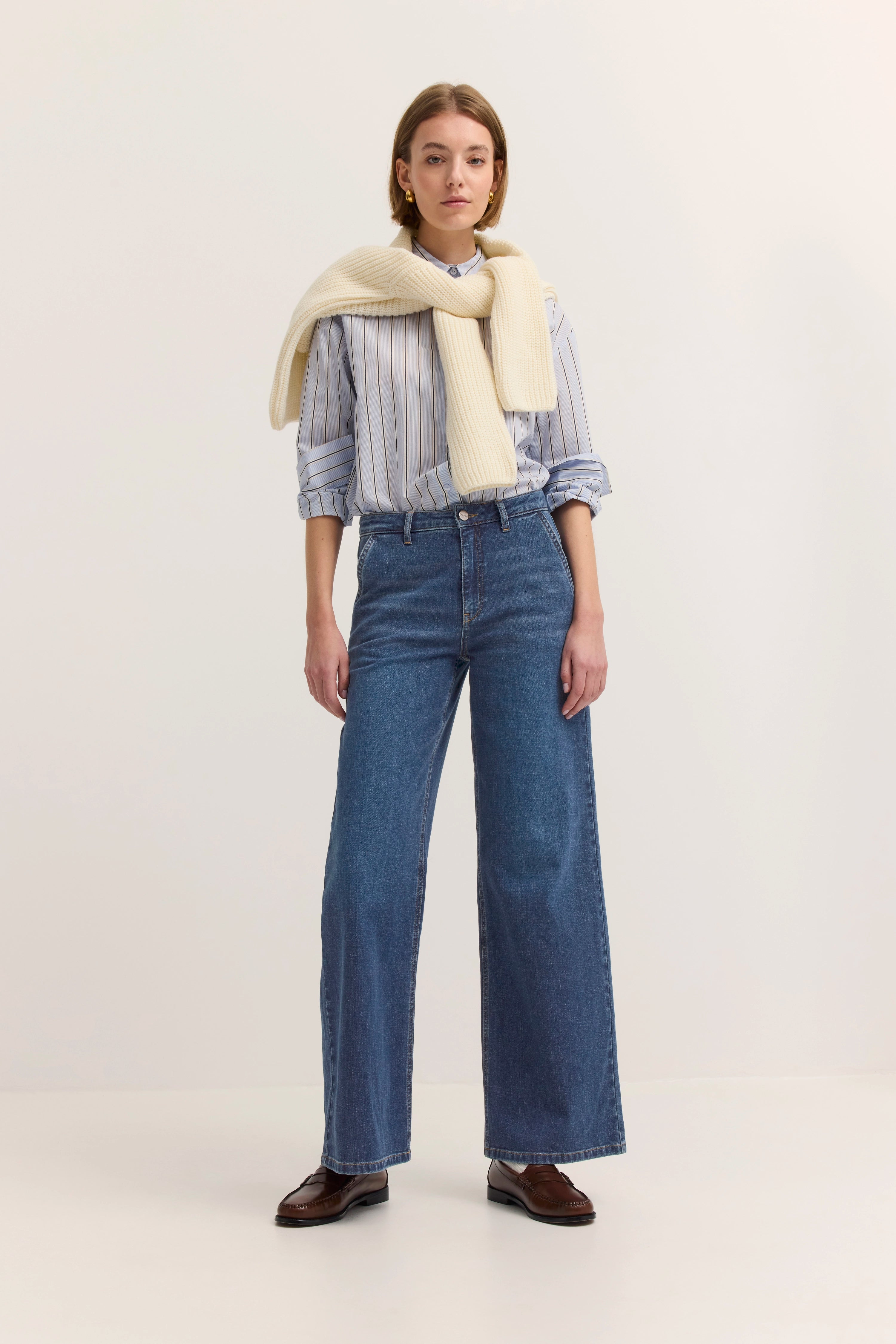 Model in blauer Jeans, gestreifter Bluse, beigem Pullover über Schultern. Stehpose, eleganter Casual-Look.
