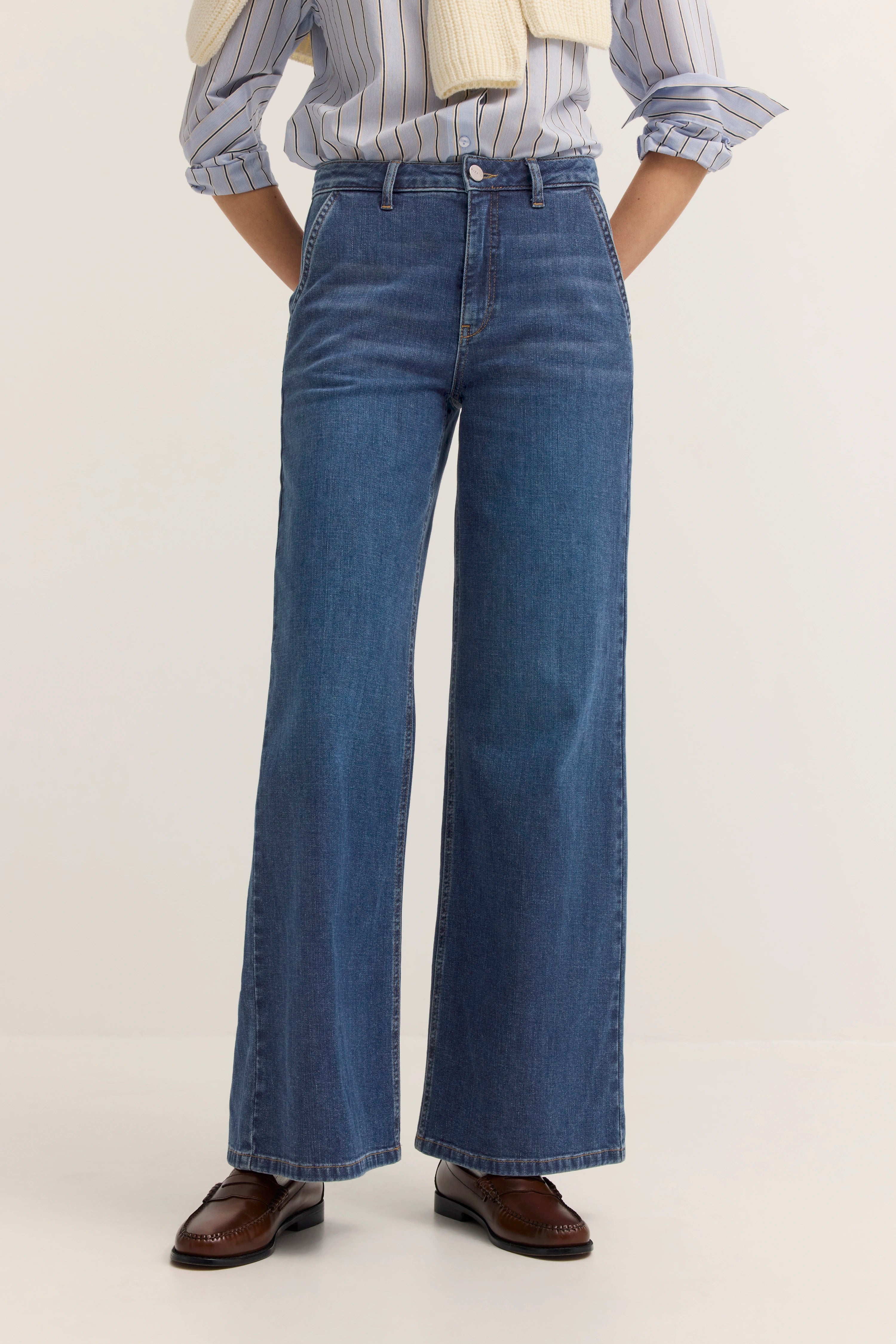 Weites Jeans-Outfit mit gestreiftem Hemd und beigefarbenem Pullover, legeres Modelbild, Arme verschränkt.