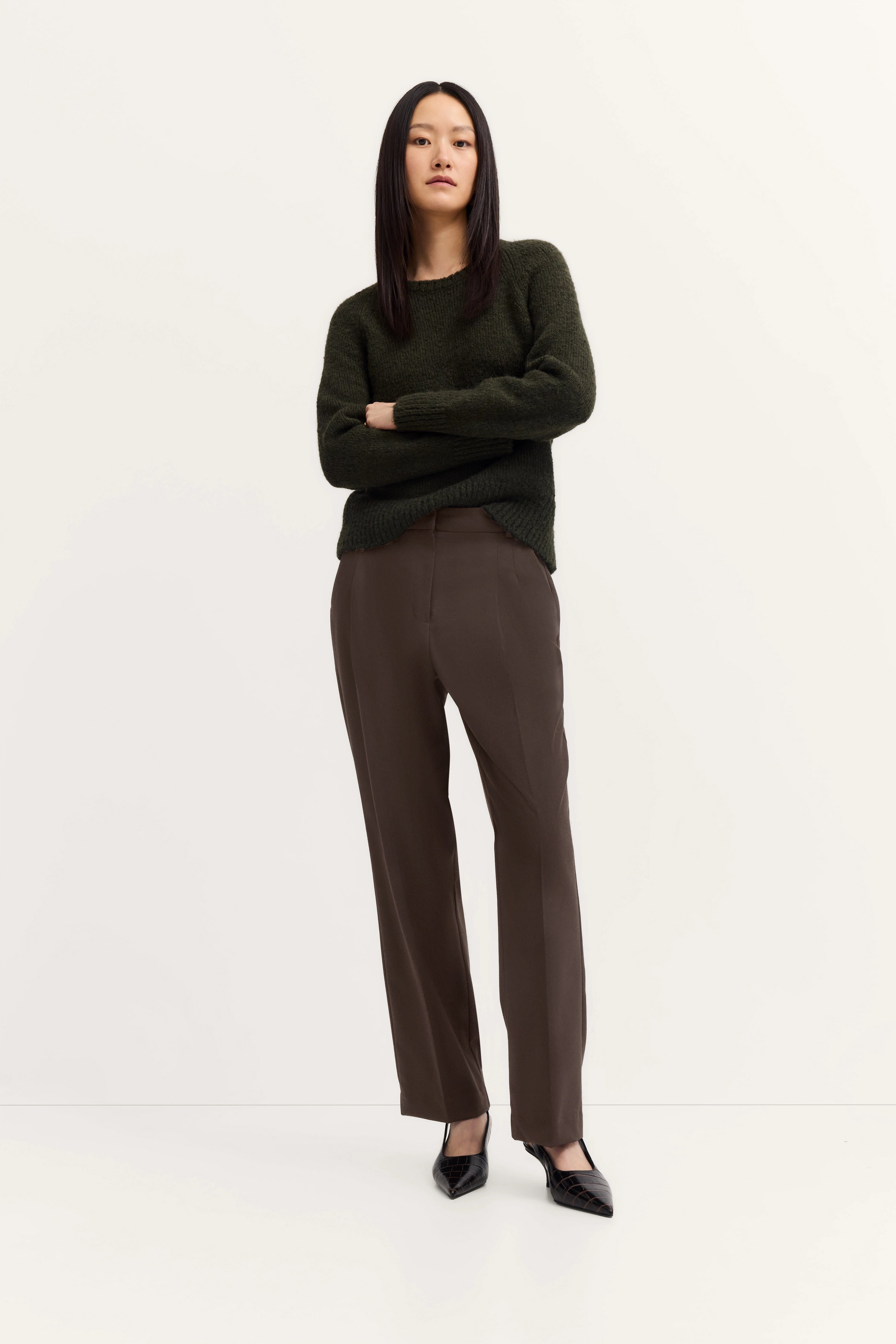 Model in dunkelgrünem Pullover und brauner Hose, Arme verschränkt, schwarze Schuhe.