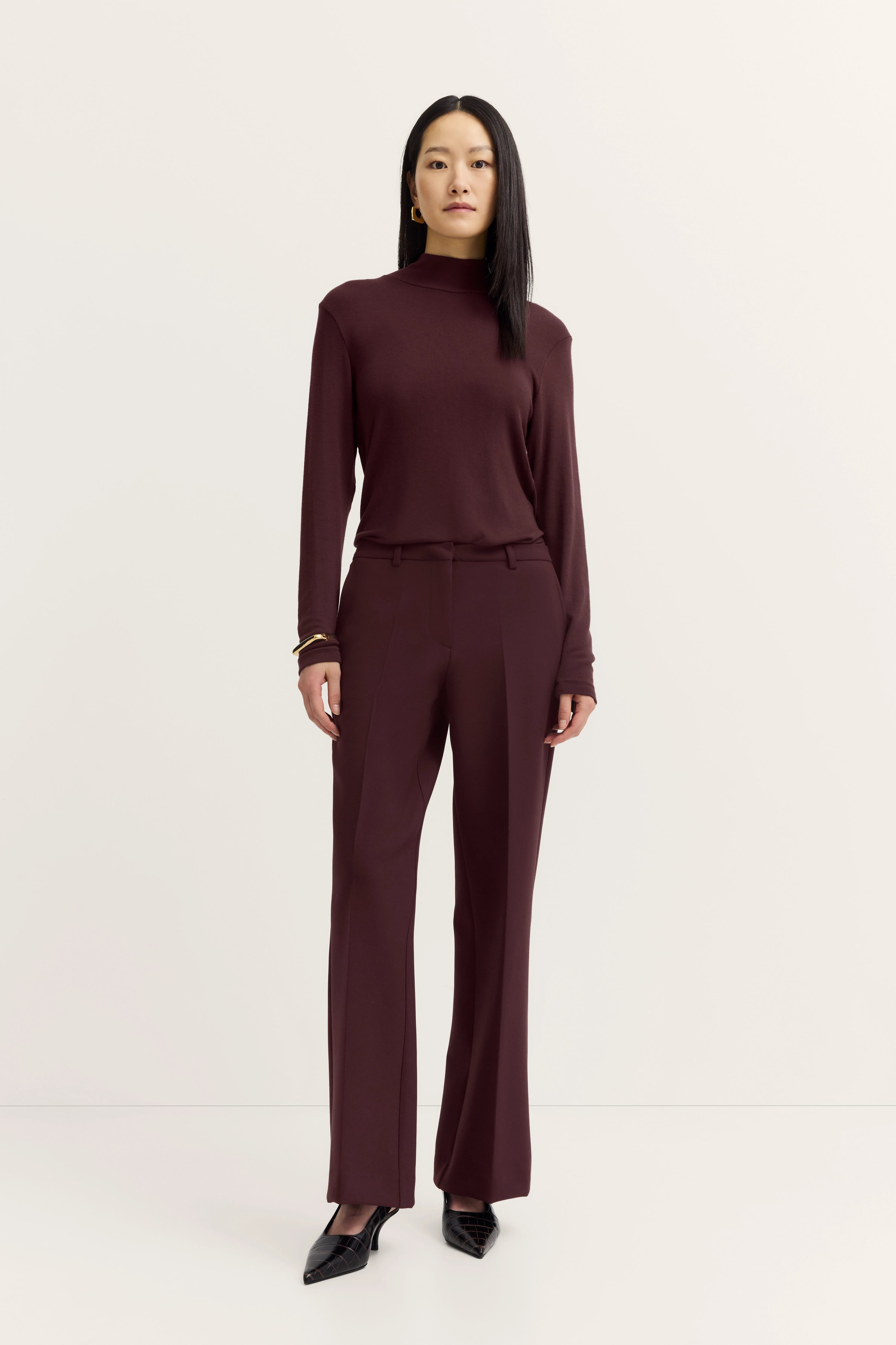 Model in bordeauxfarbenem bugatti Outfit: Rollkragenpullover, Hose, schwarze Schuhe, stehende Pose.