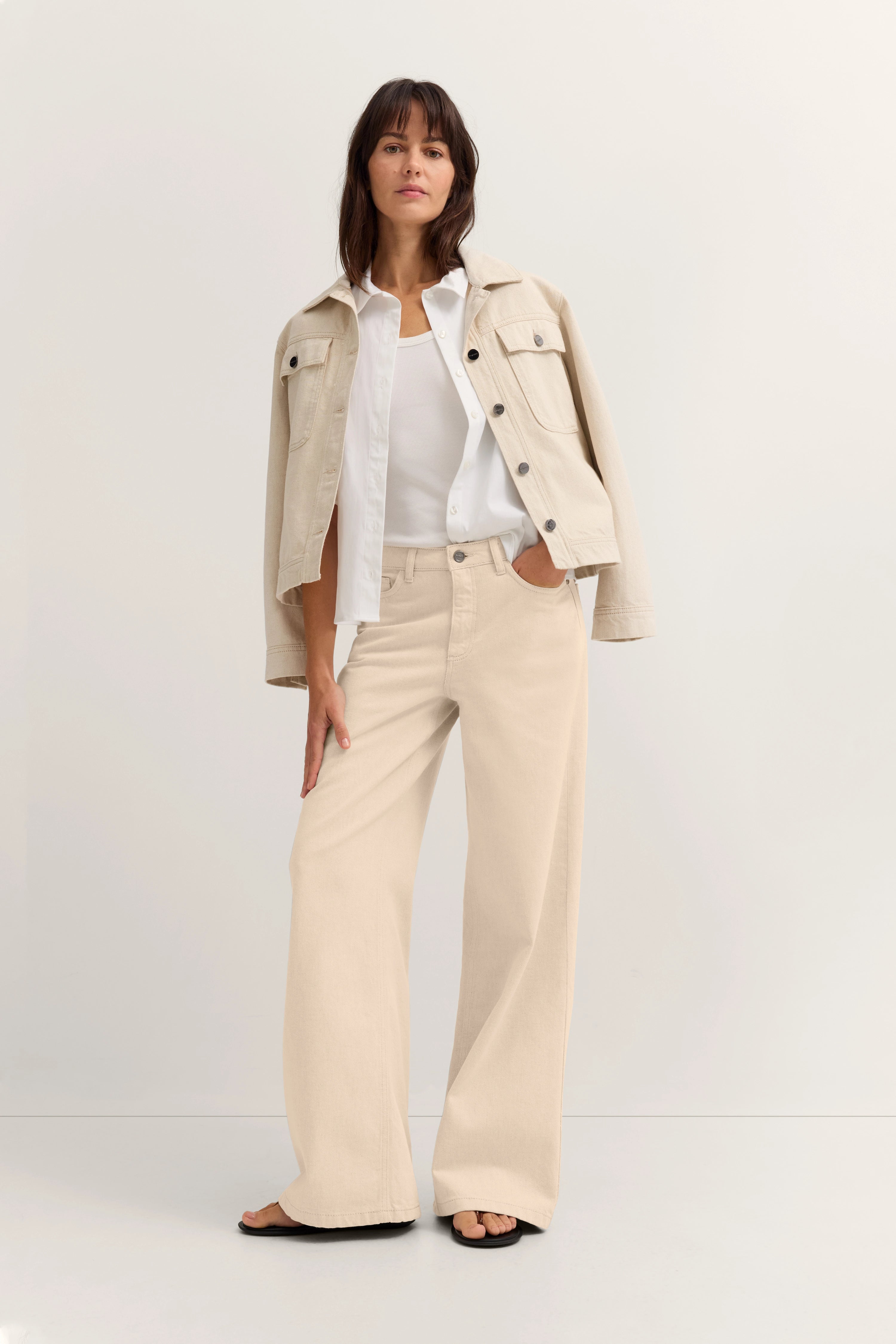 Model in beigefarbener Jacke, weißem Hemd und Top, weiten beigefarbenen Hosen, steht lässig.