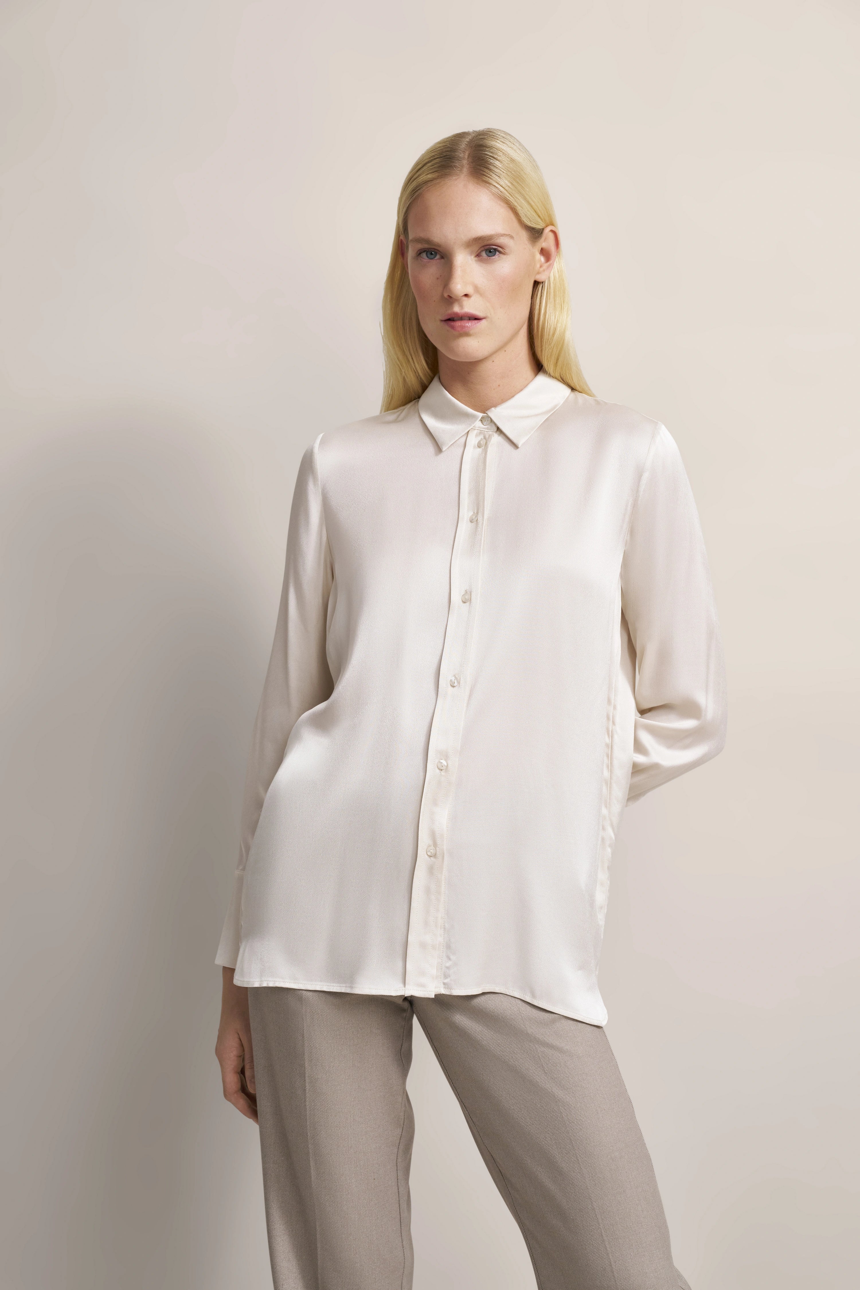 Elegante bugatti Bluse in Creme, kombiniert mit beigen Hosen. Modelbild, lässige Pose, Hände in Taschen.