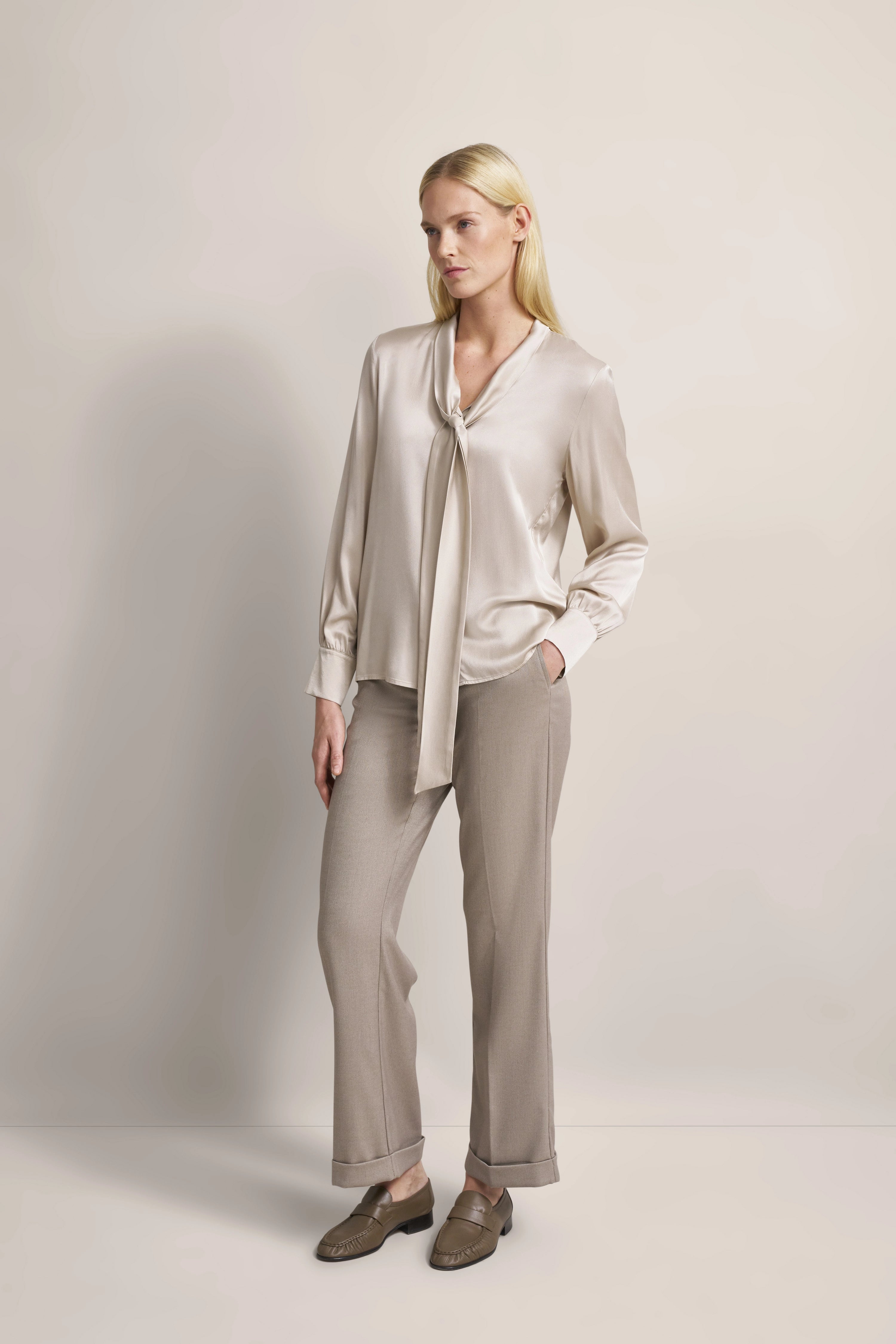 Elegante Modelaufnahme mit beiger Bluse, lockerem Schnitt und beiger Hose. Entspanntes, seitliches Posing.