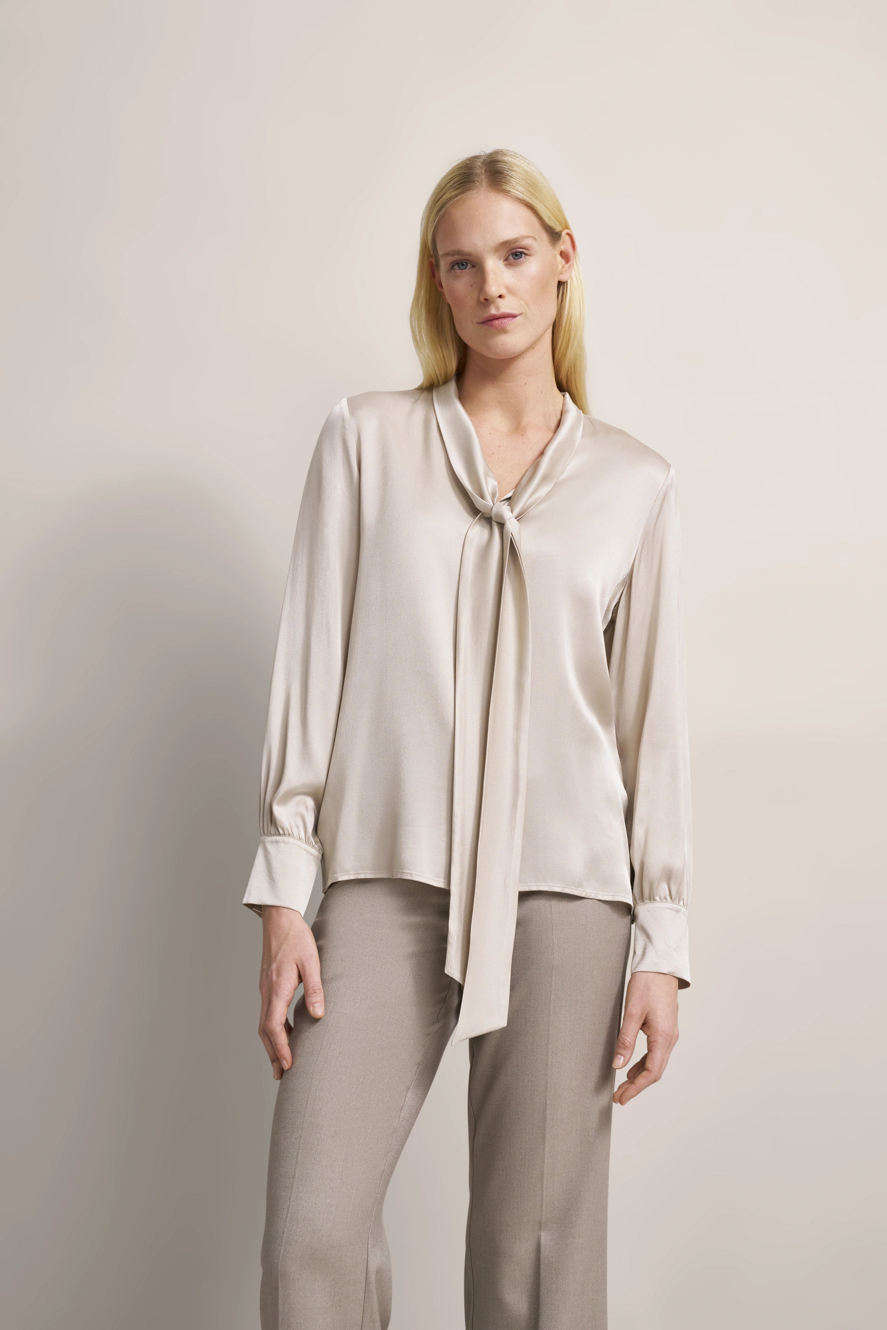 Elegantes Modelbild mit beiger Seidenbluse mit Krawattenkragen und grauer Hose, leichtes Posing.