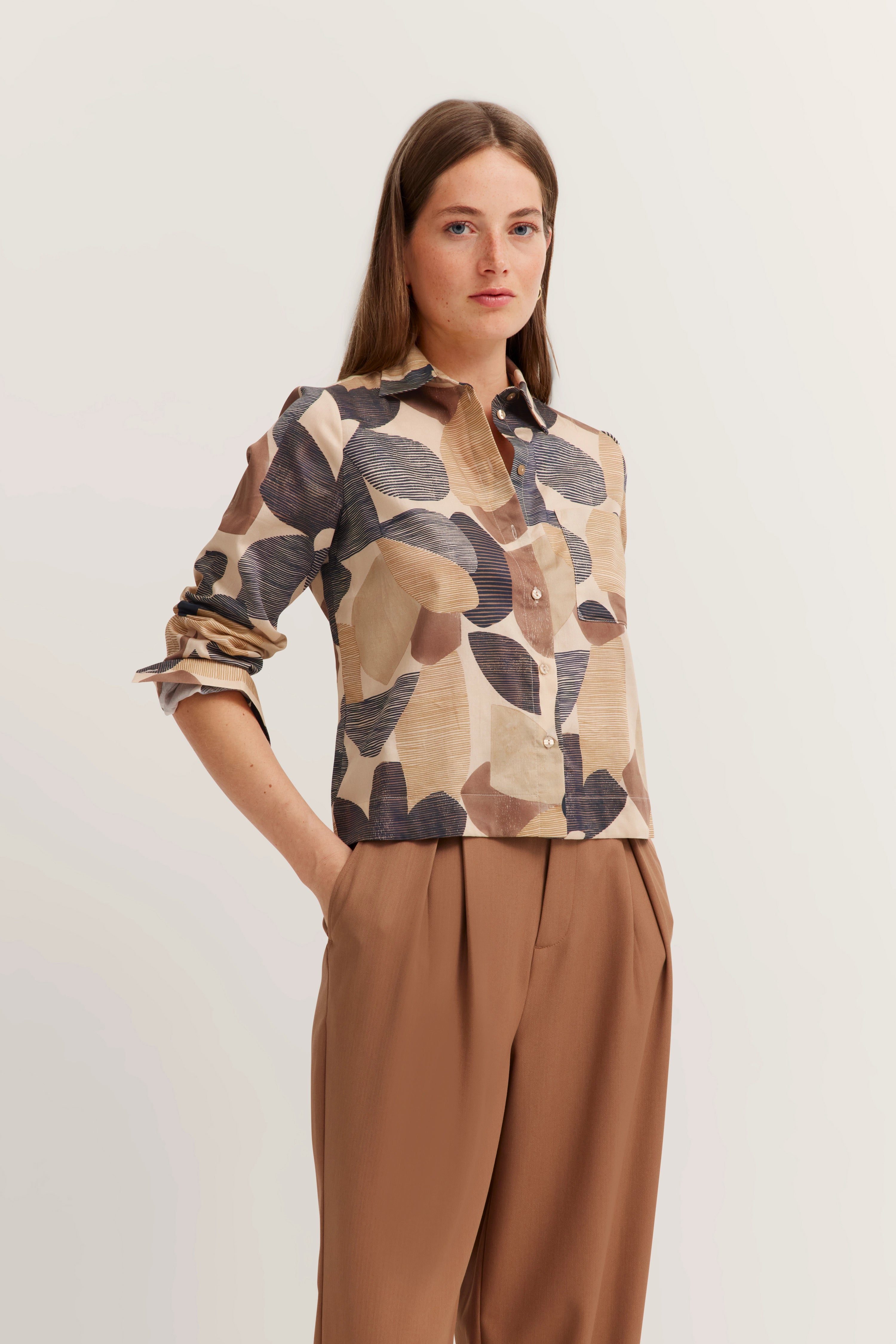Bluse mit geometrischem Muster in Beige und Braun, locker gestylt, Model stehend, eine Hand in der Tasche.