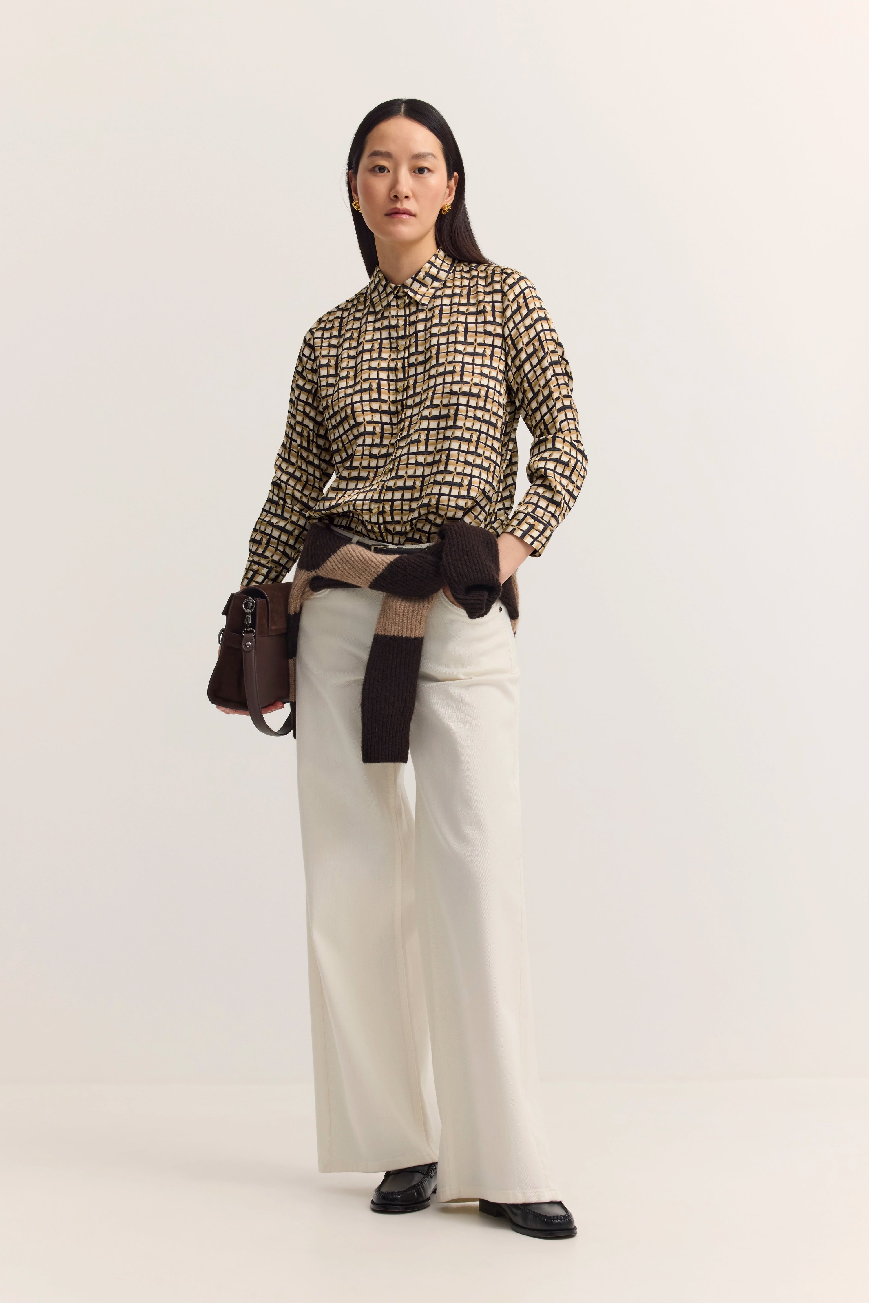 Model in gemusterter Bluse, weißer Hose, brauner Pullover um Hüfte, lässige Pose.