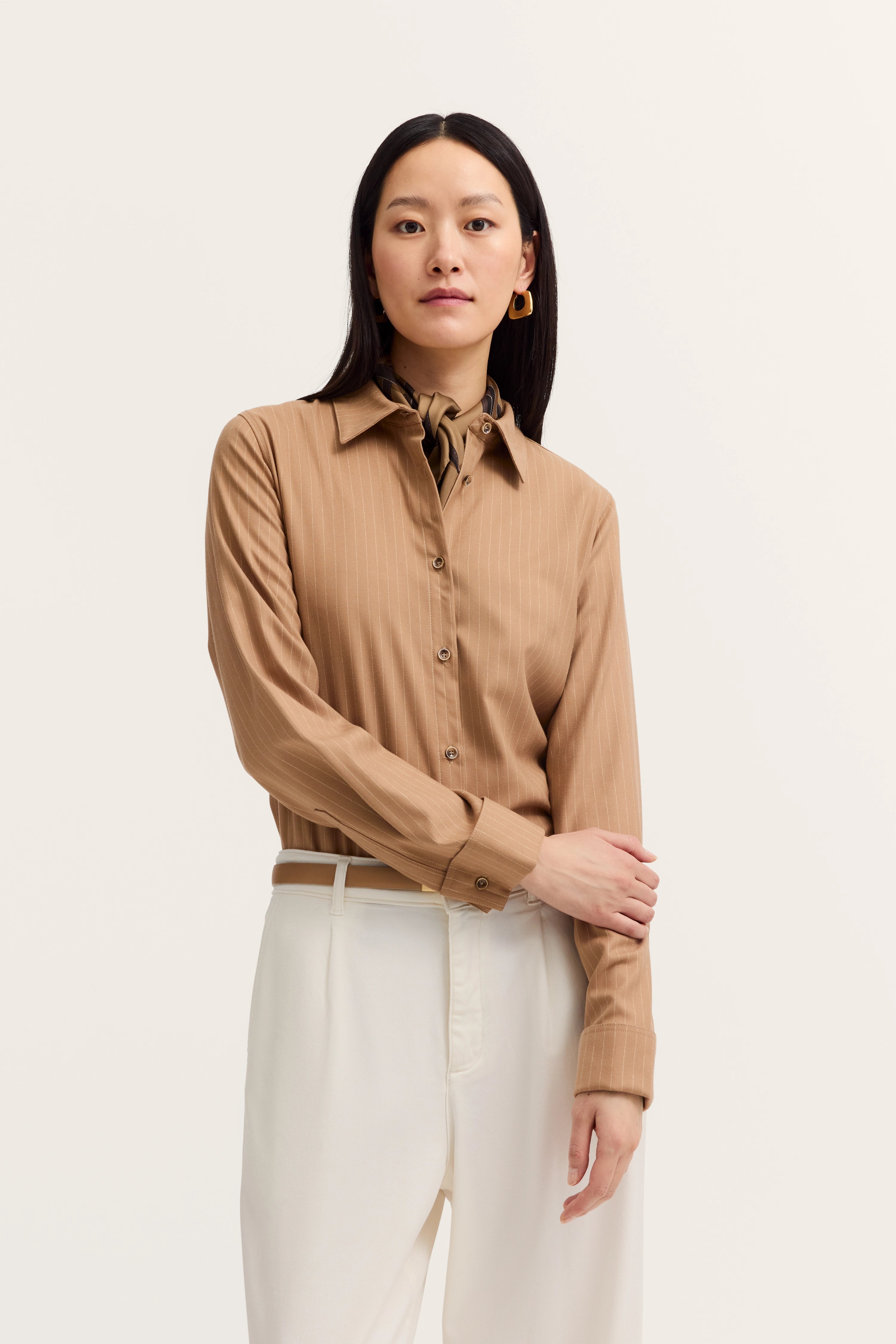 Elegantes Model in beige gestreifter Bluse mit weißer Hose von bugatti, Arme verschränkt.