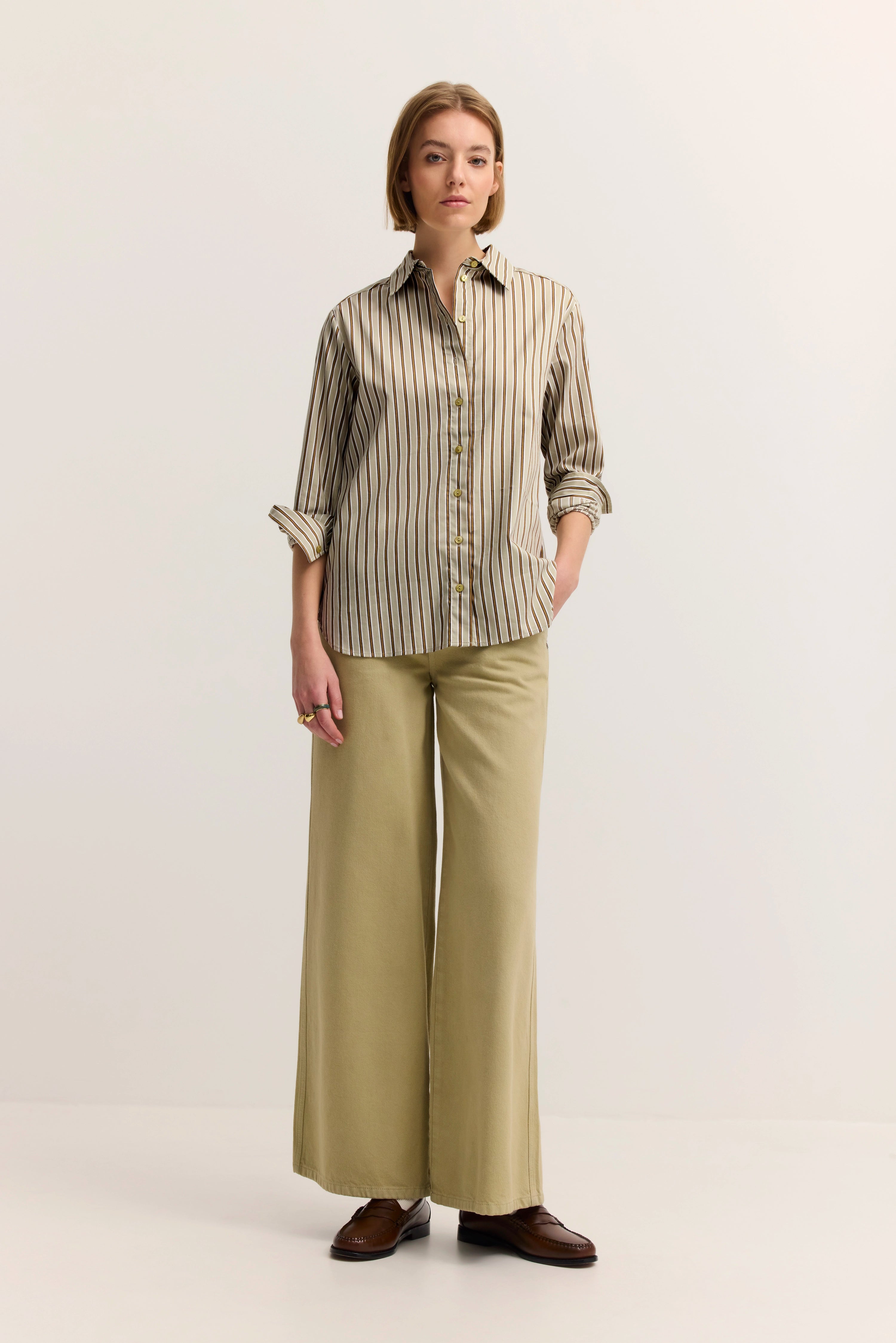 Gestreiftes Hemd in Beige, weite Hose in Khaki, braune Schuhe, Model steht locker, Hände in Taschen.