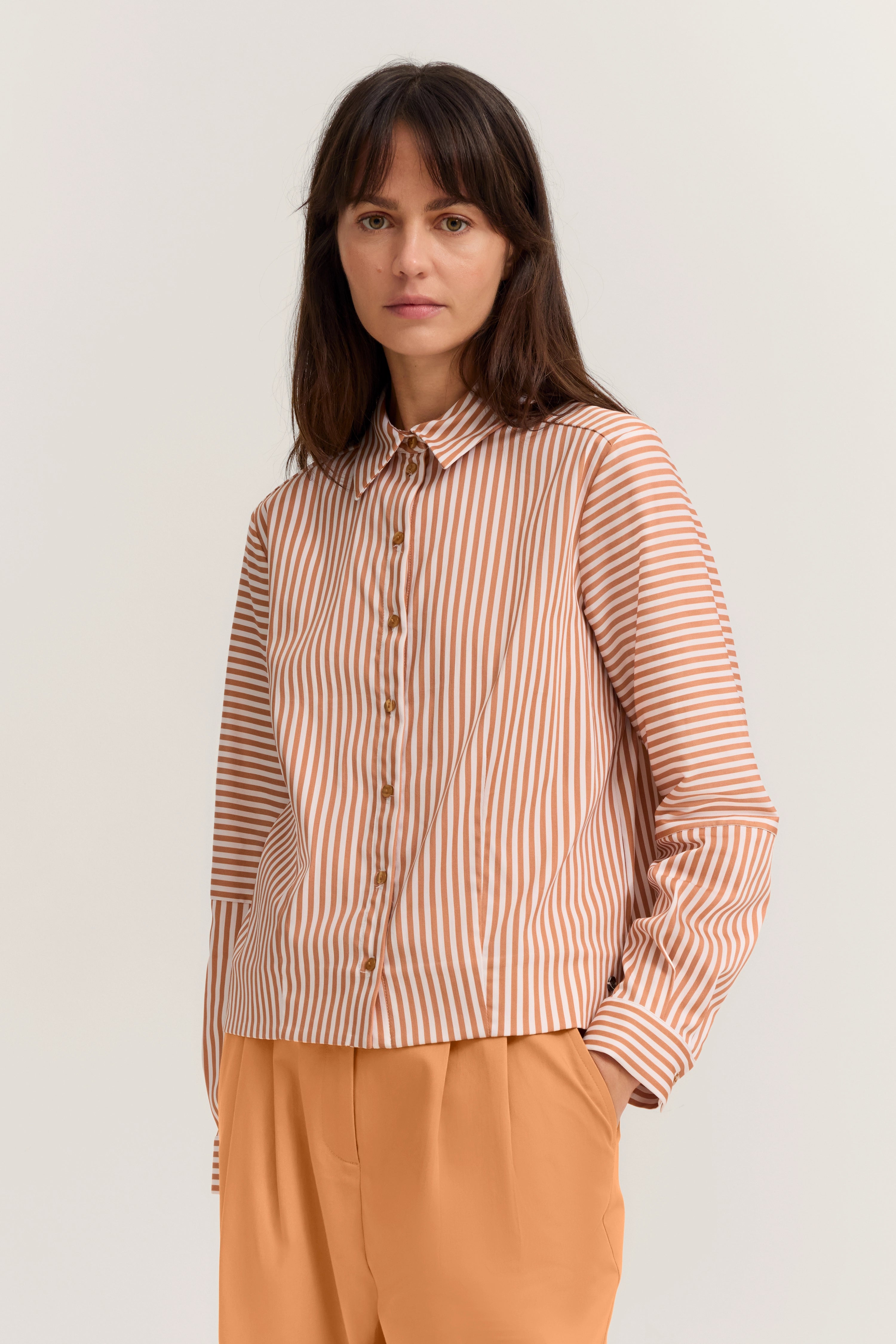Streifenbluse in Beige-Weiß auf Model, kombiniert mit beigefarbenen Hosen, legeres Posing.