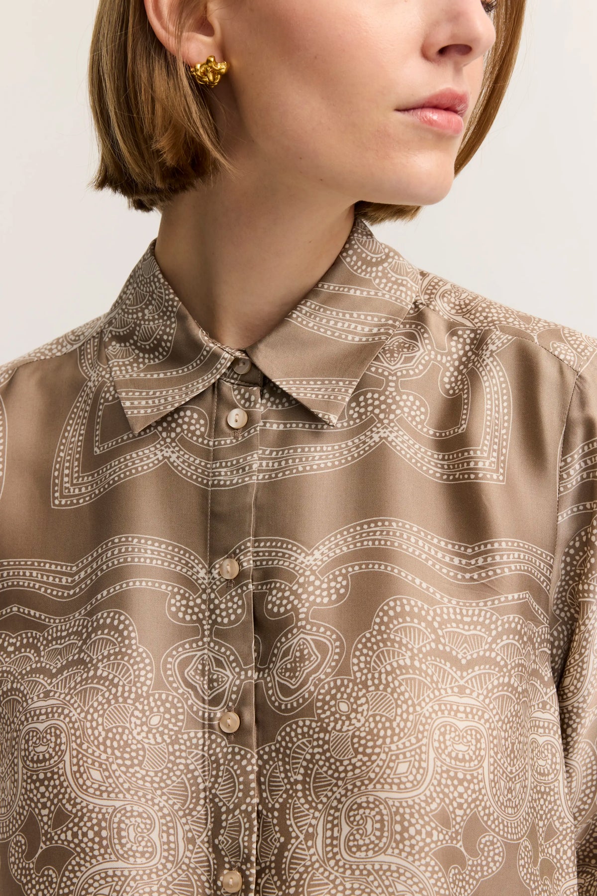 Seidenbluse in Beige mit weißem Paisley-Muster, Büstenbild, goldene Ohrringe.