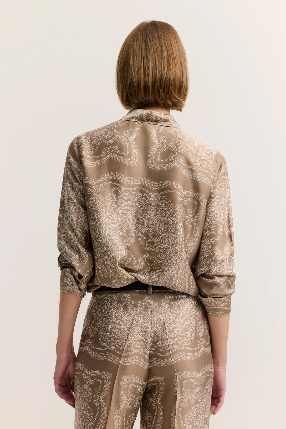 Beige gemustertes Outfit mit langer Bluse und Hose, Model von hinten, entspanntes Posing.