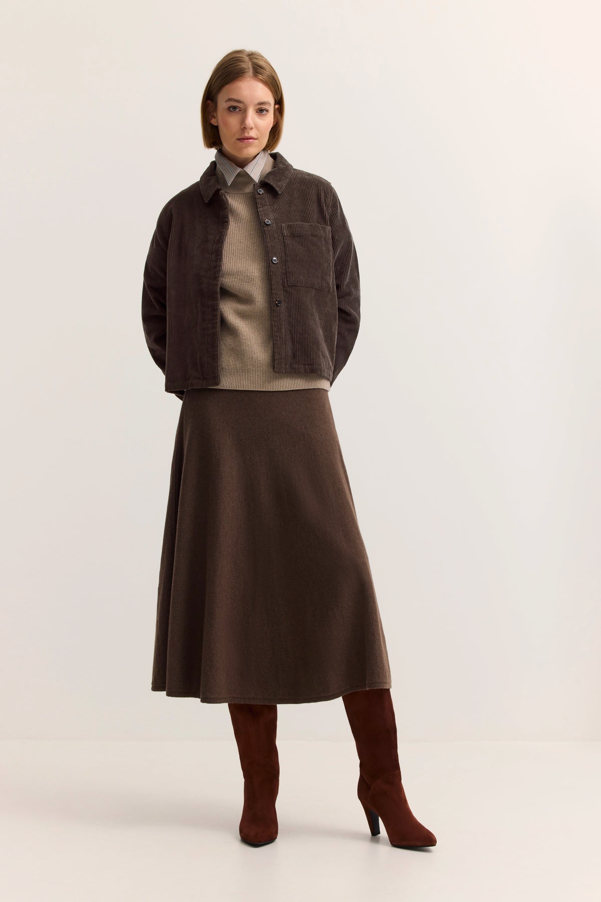 Model trägt braune Jacke, beigen Pullover, braunen Rock und Stiefel. Stehende Pose.