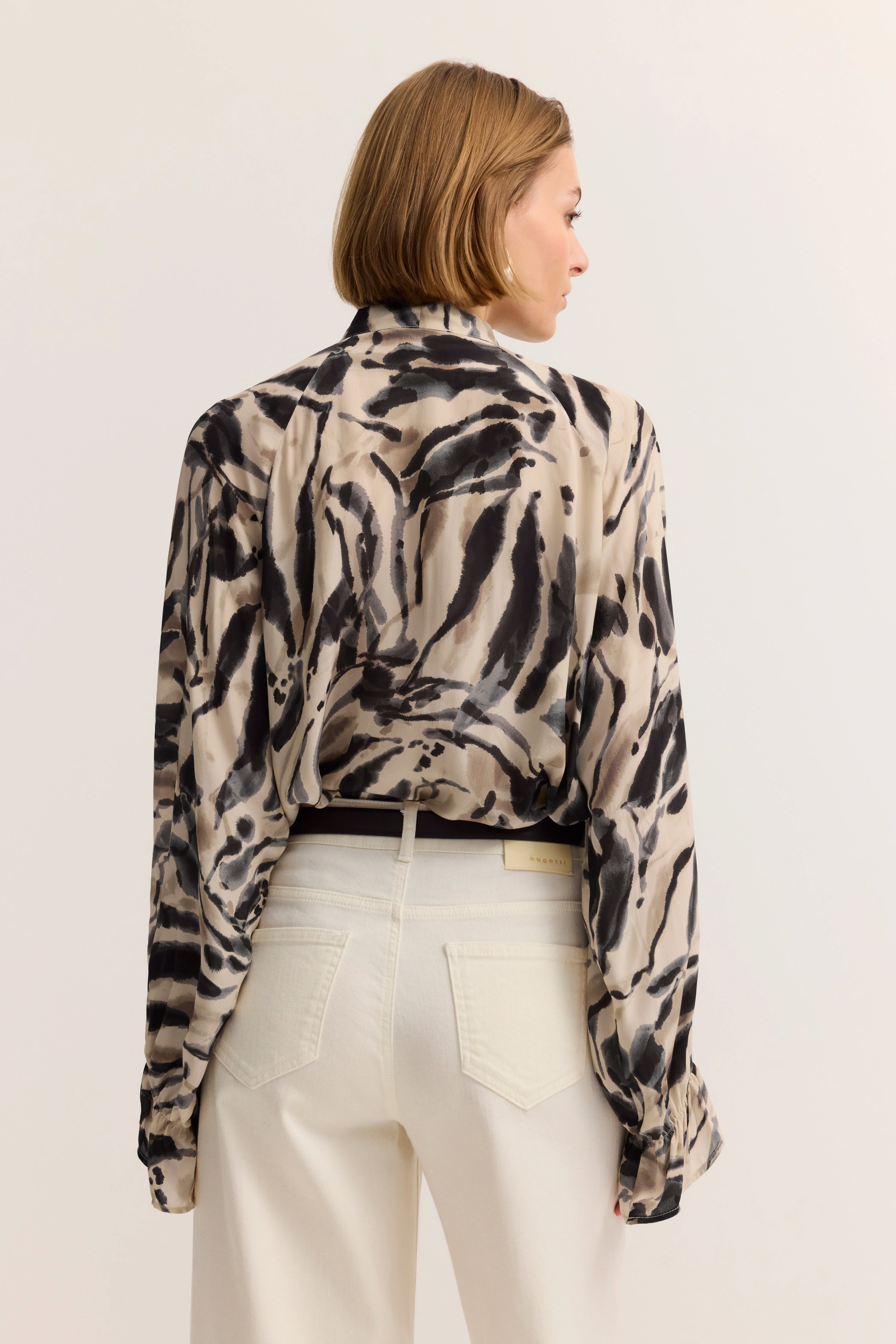 Beige-schwarze gemusterte Bluse mit weißer Hose, Model von hinten, entspannte Haltung.