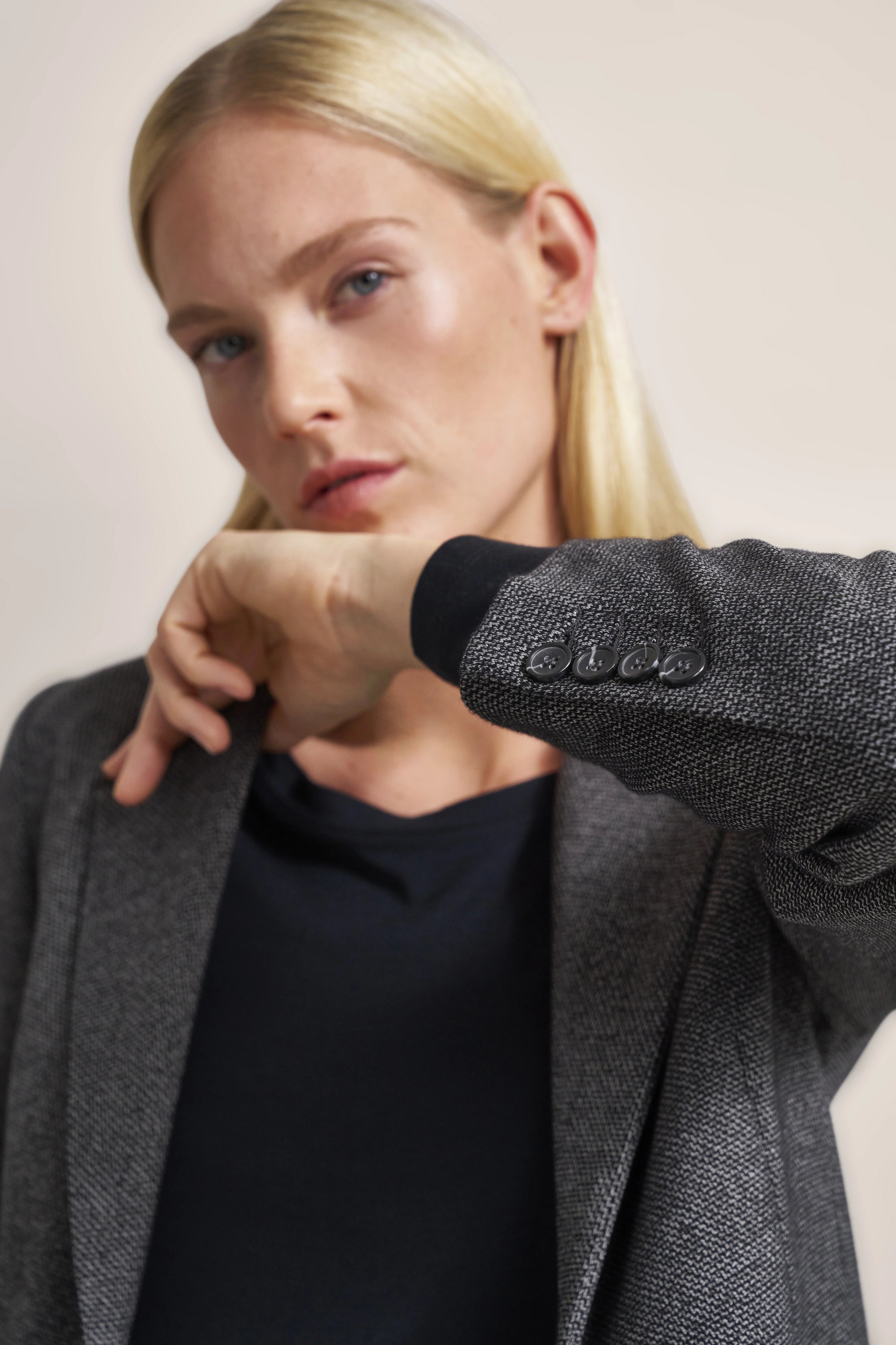 Elegantes Model im grauen bugatti Blazer, schwarzes Shirt, lässige Pose mit Hand am Kinn.