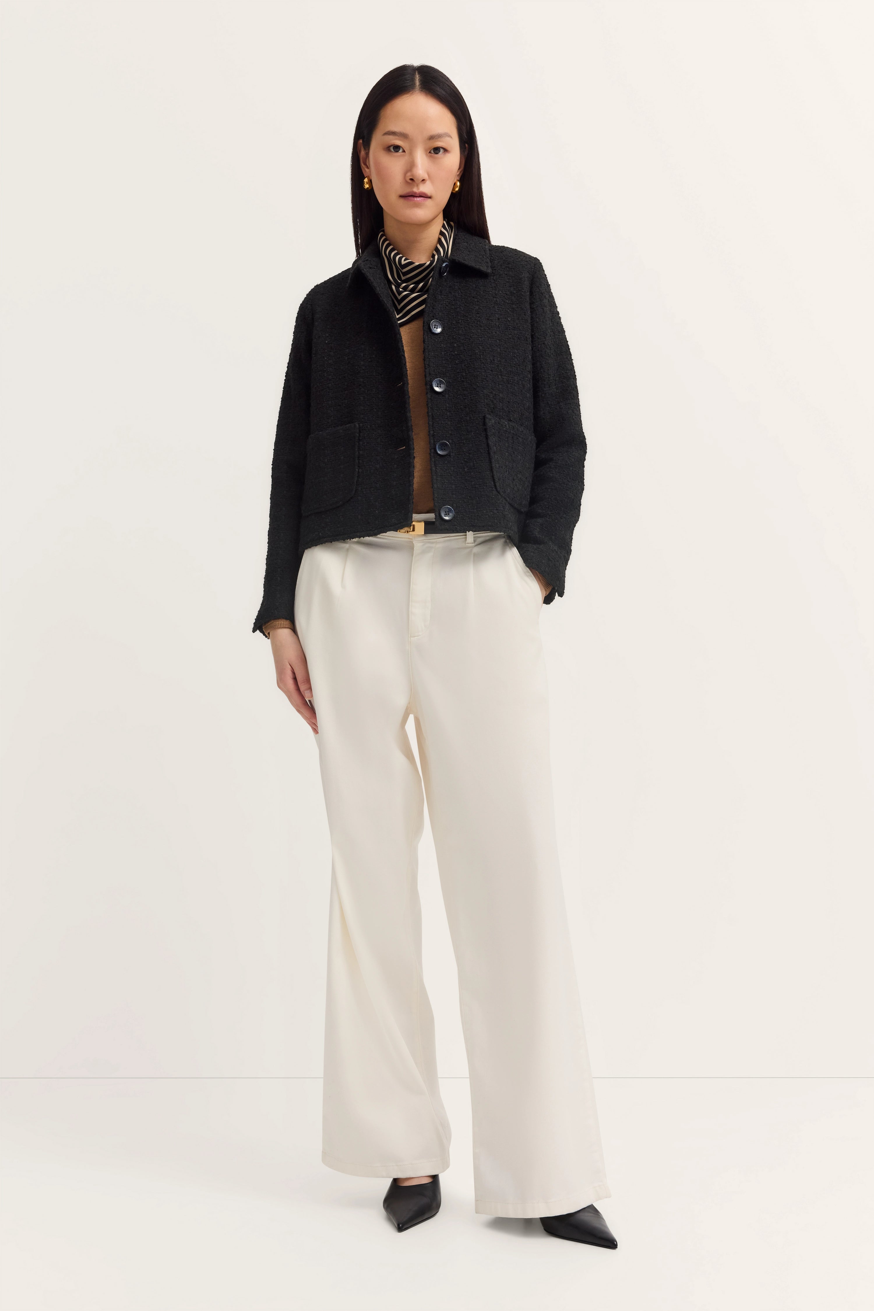 Model: Zwarte jas, beige trui, gestreept shirt, witte broek. Casual met handen in de zakken.