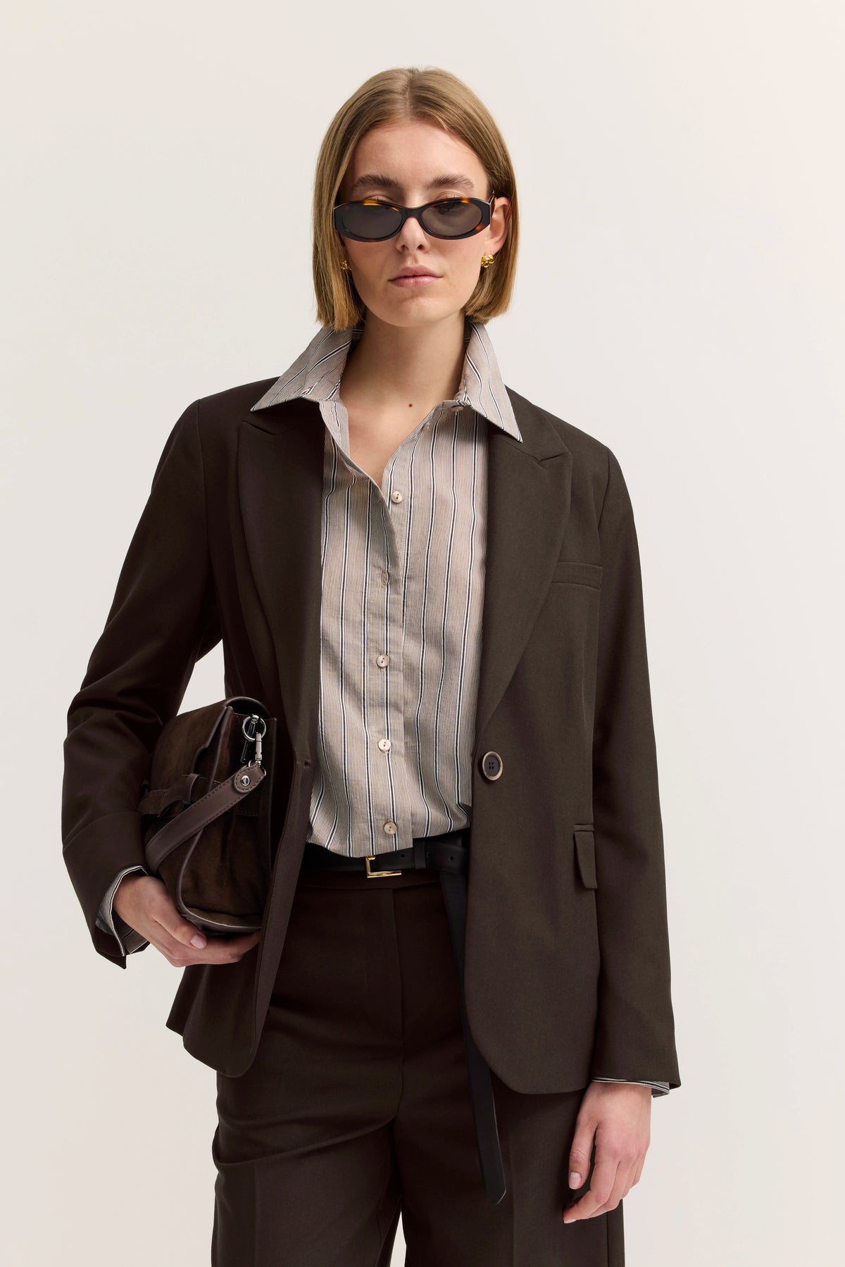Elegantes Model in braunem bugatti-Anzug, gestreiftem Hemd, posiert selbstbewusst mit Sonnenbrille.