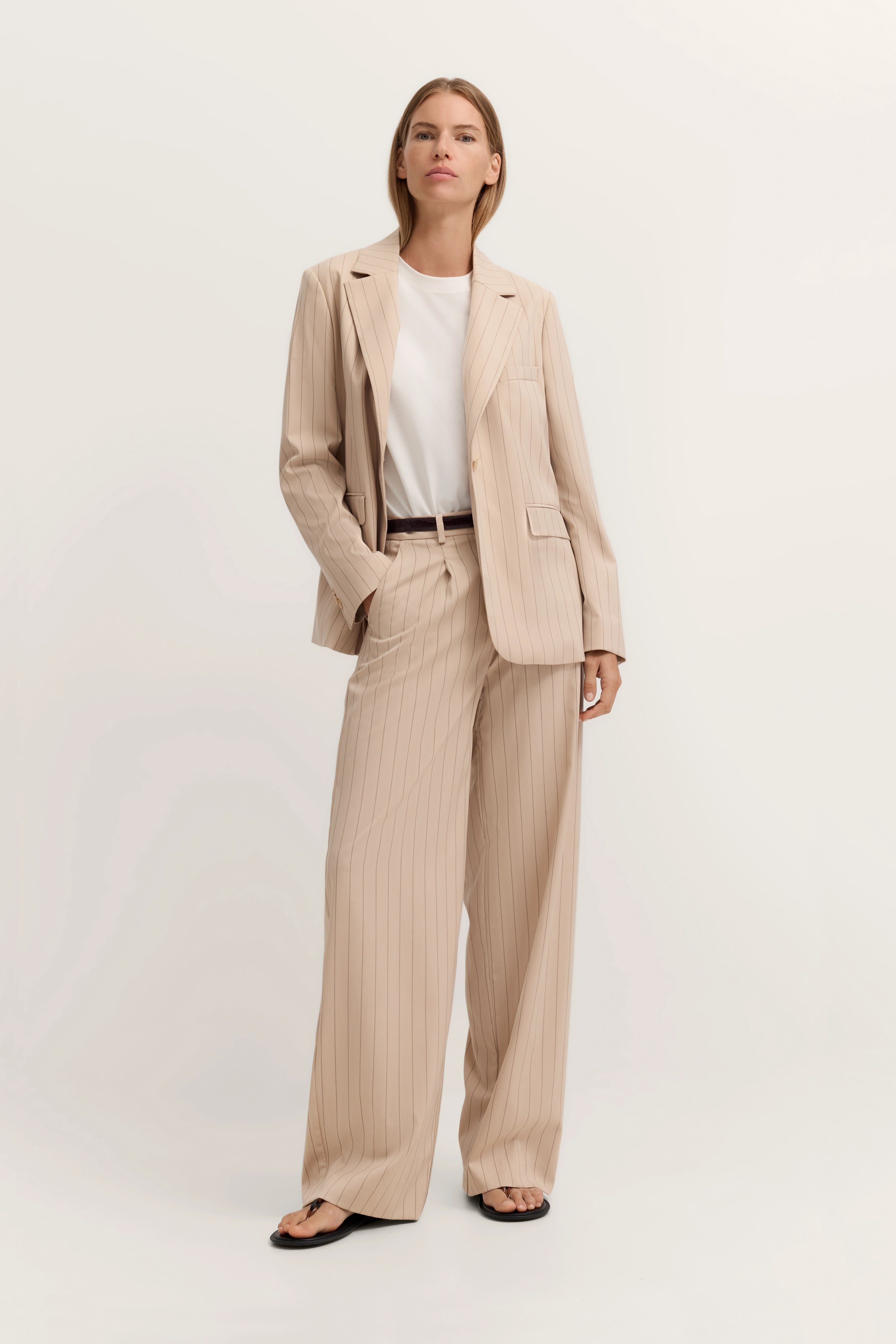 Beigefarbener Nadelstreifenanzug mit weißem Shirt, Model stehend mit Händen in den Taschen.