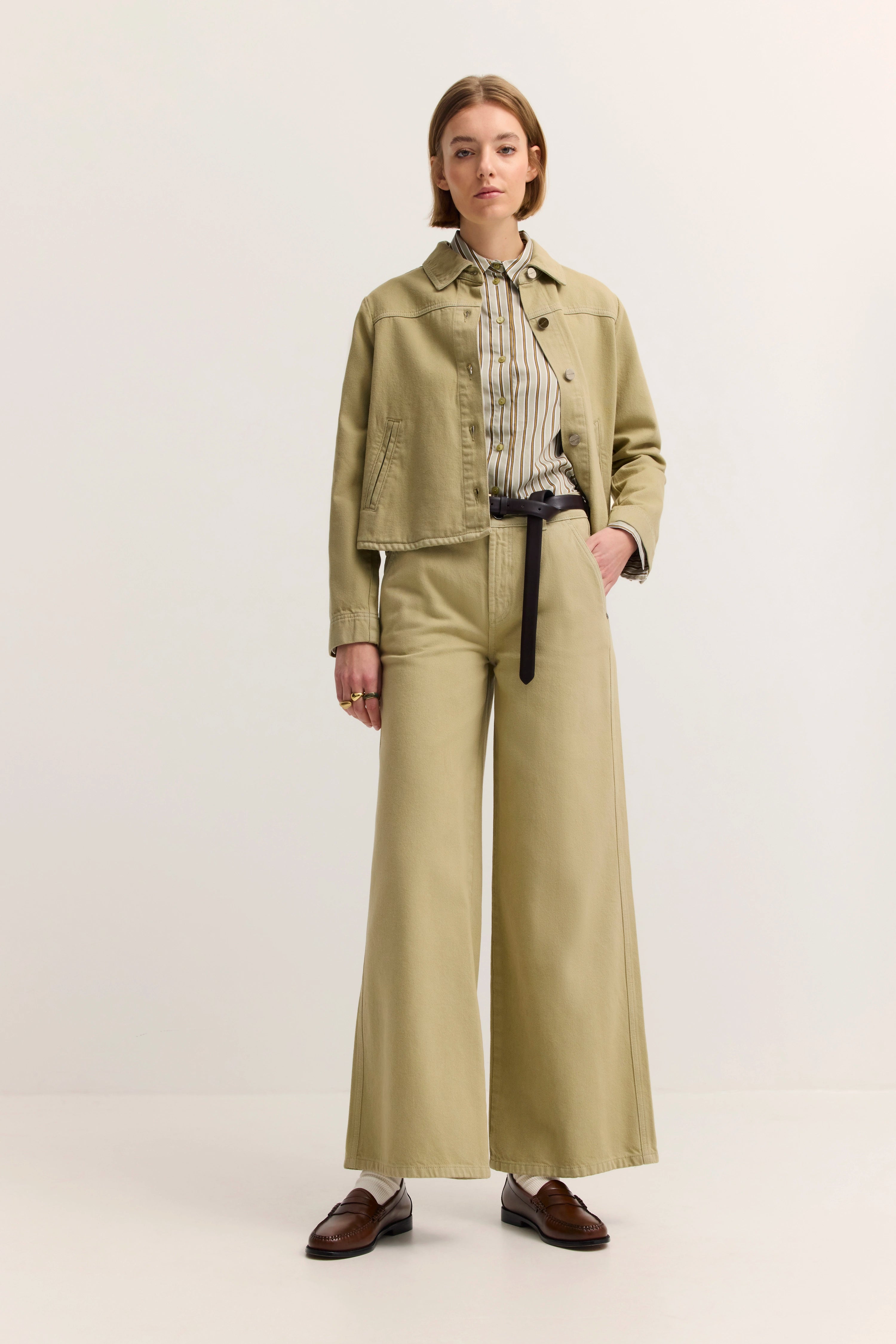 Model in sandfarbenem Jackenset und gestreiftem Hemd, schwarze Gürtel, braune Schuhe.