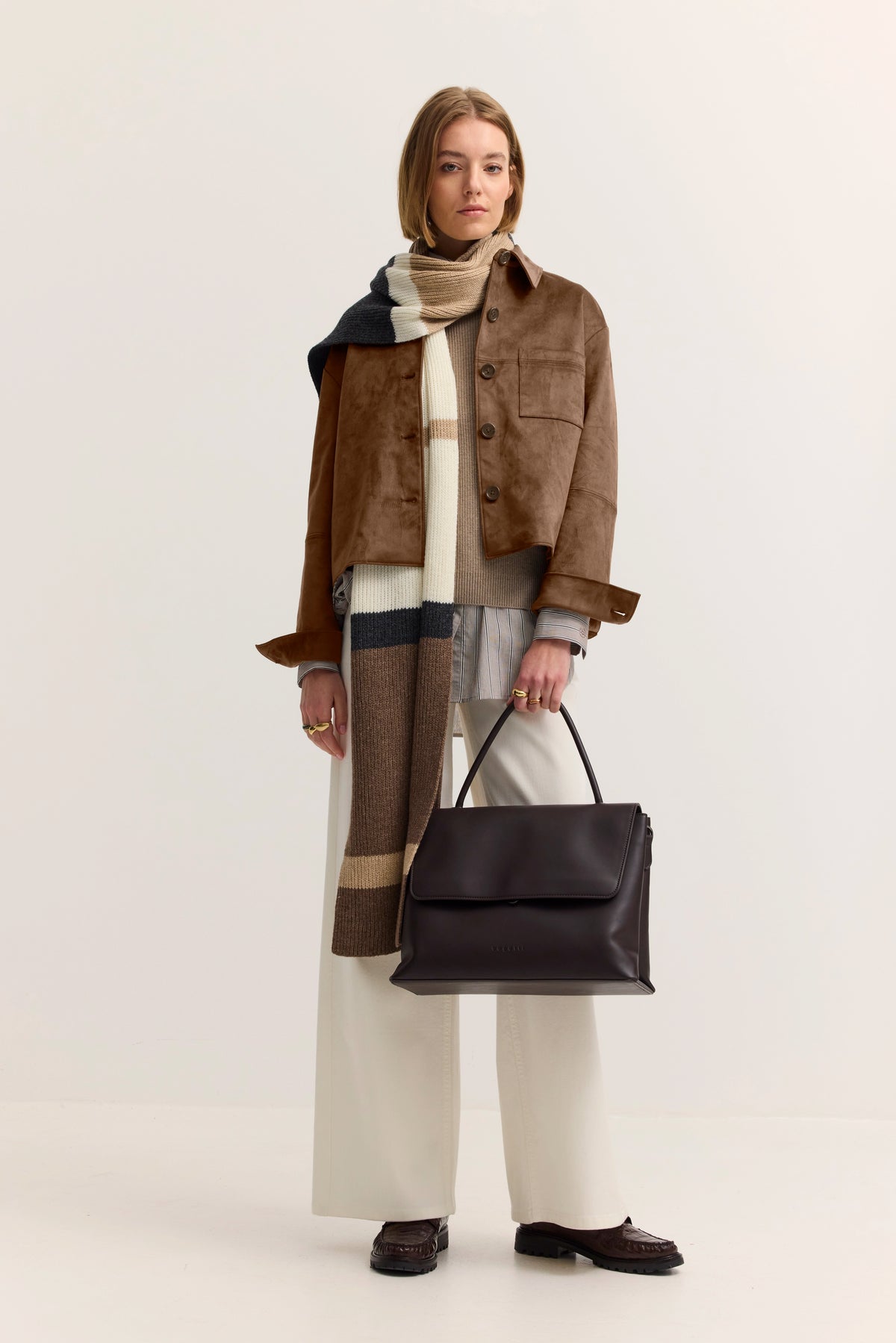 Model trägt braune Jacke, gestreiften Schal, weiße Hose
