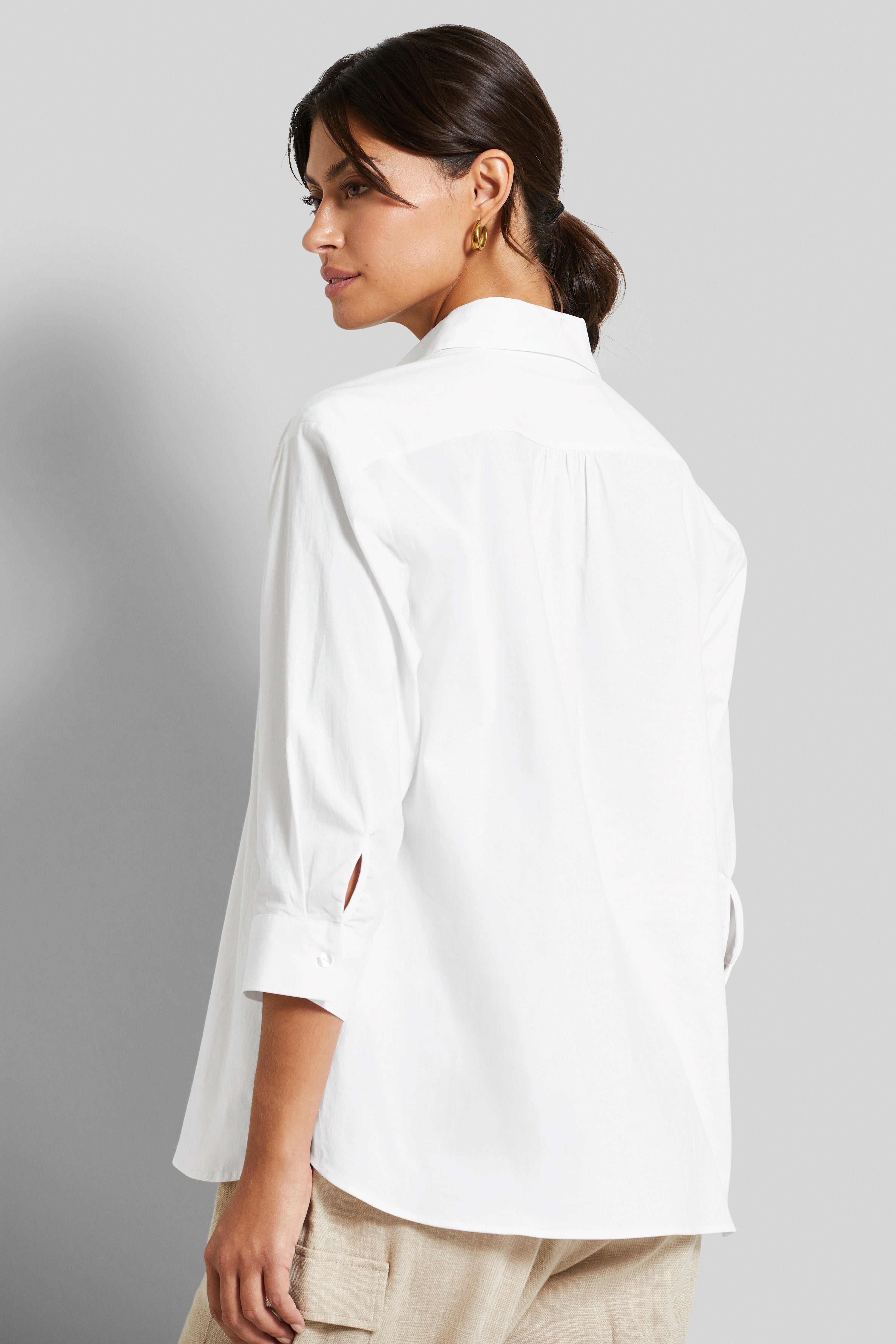 Weiße Bluse mit 3/4-Ärmeln, Beige Hose, Model legt entspannt die Hand an die Seite.