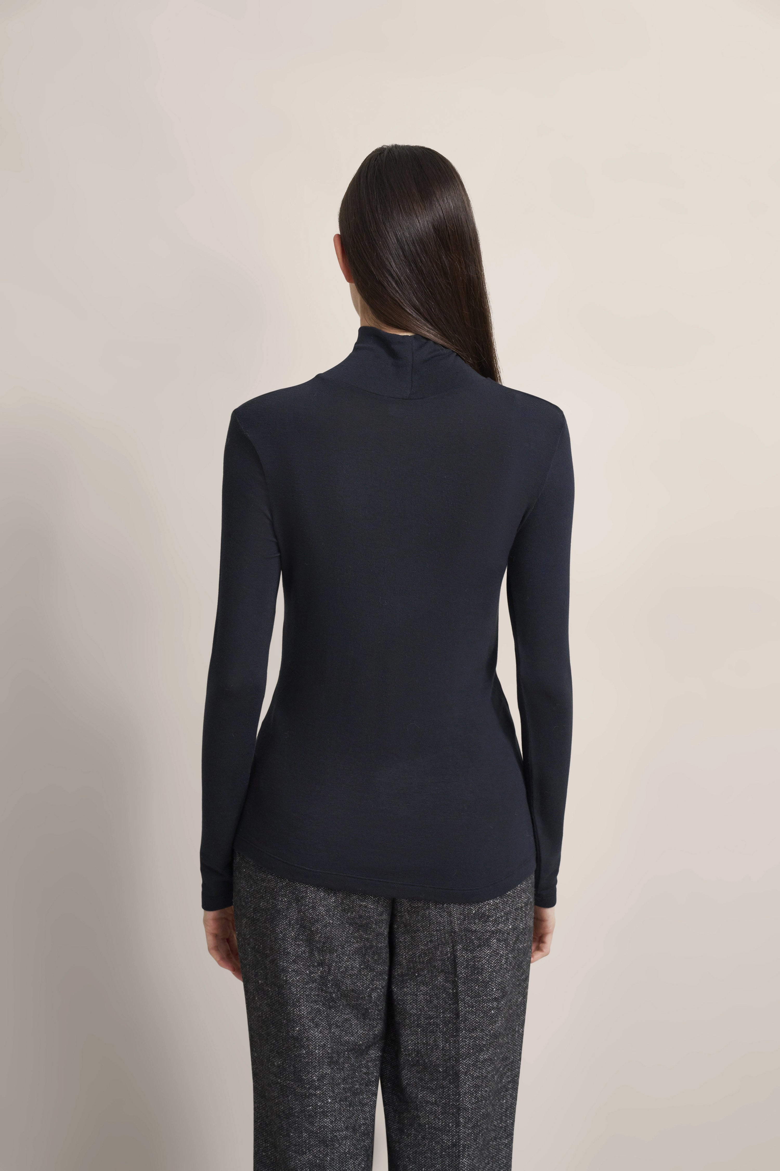 bugatti Modelbild: Frau trägt dunkelblauen Rollkragenpullover und graue Hose, Rückansicht, entspannte Haltung.
