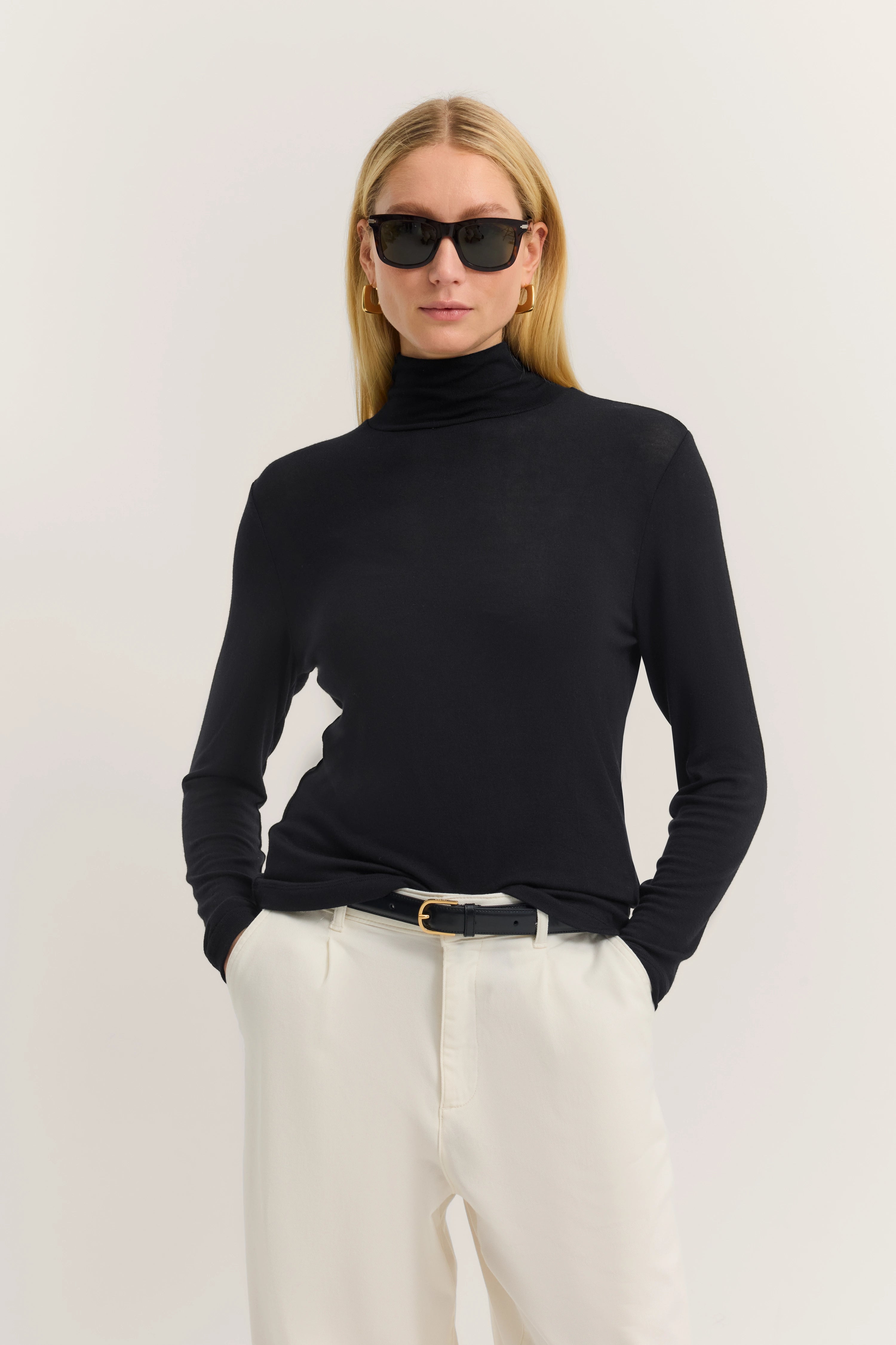 Schwarzer Rollkragenpullover, weiße Hose, Model mit Sonnenbrille, lässige Pose.