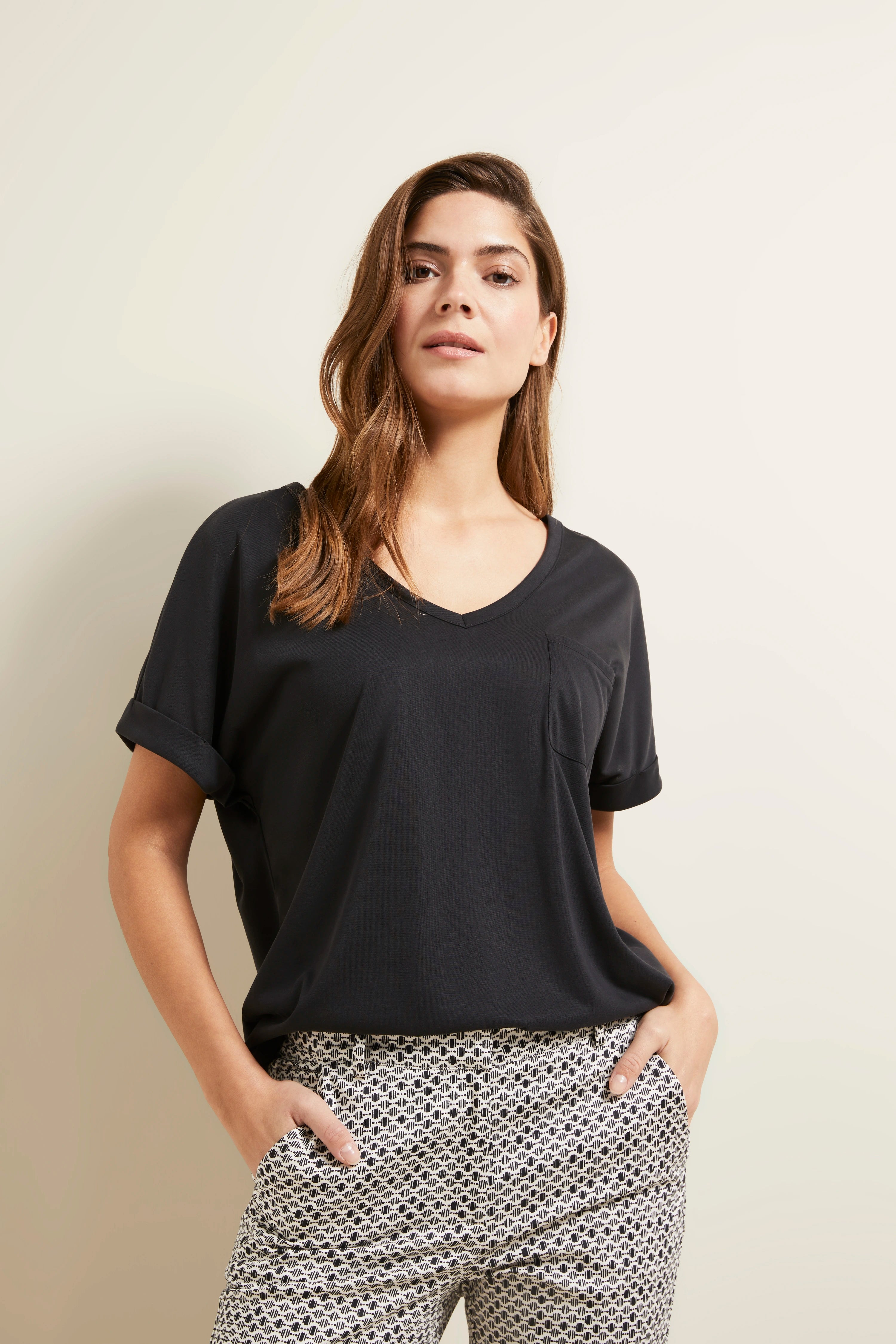 Modelbild: Frau trägt schwarzes V-Ausschnitt-Shirt und gemusterte Hose. Hände in Taschen, entspannte Pose.