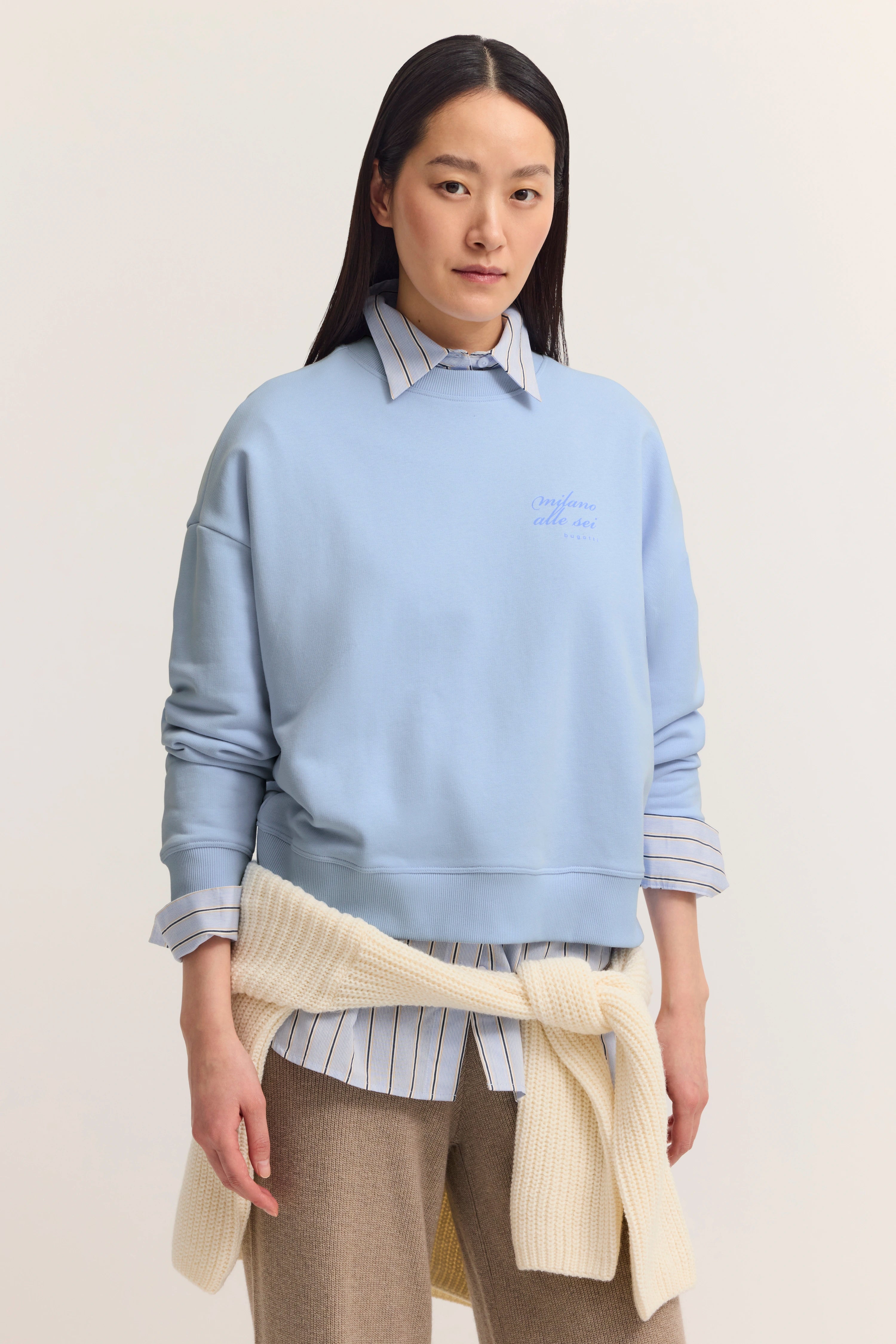 Model trägt hellblauen bugatti Pullover über gestreiftem Hemd, cremefarbener Strick um Hüfte gebunden.