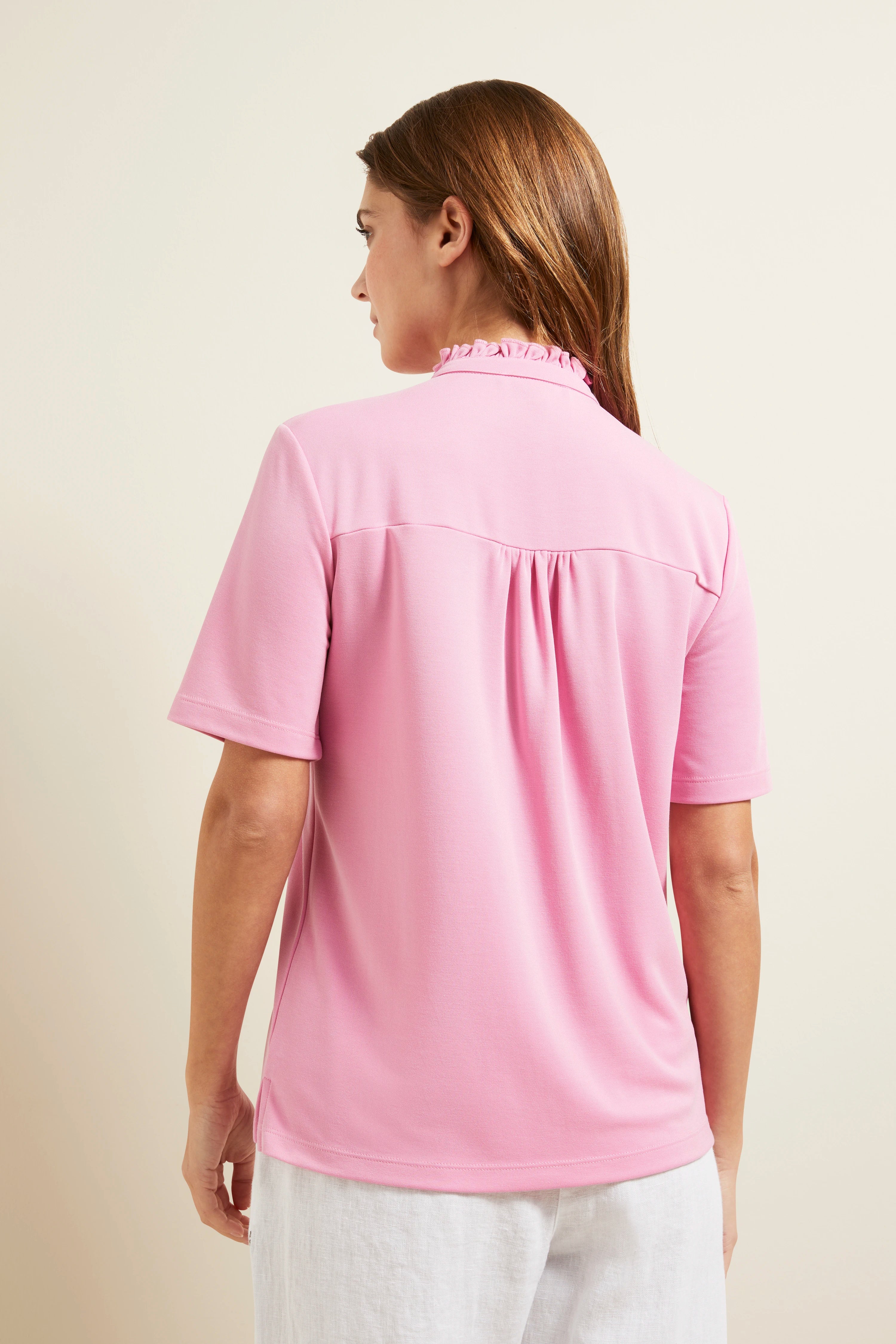 Rugaanzicht van vrouw in roze bugatti shirt, ontspannen stijl, witte broeksband zichtbaar.