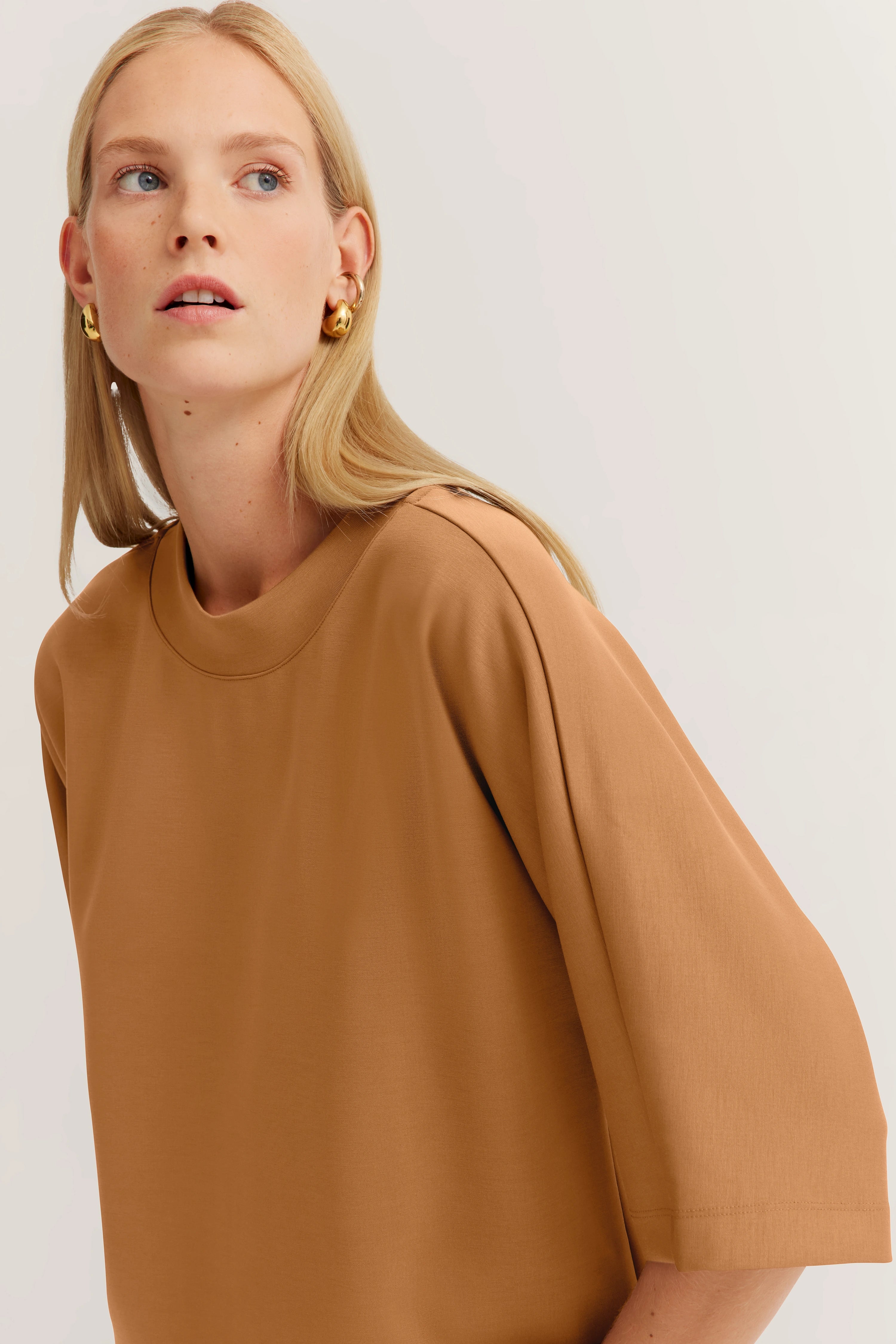 Model in camelkleurige top, ontspannen pose, elegante oorbellen.