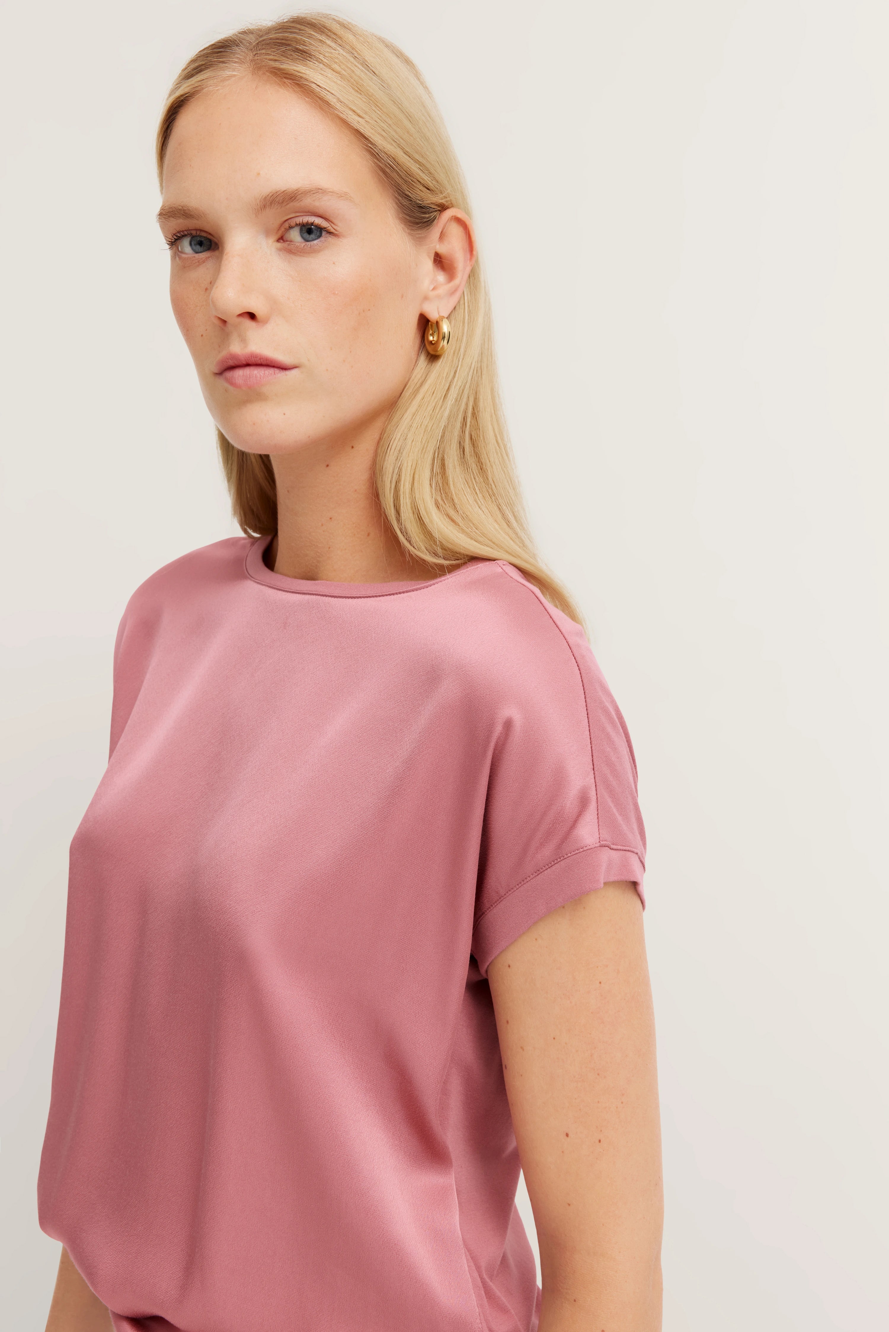 Model in roze bugatti-shirt, casual look, zijwaartse pose, gouden oorbellen.