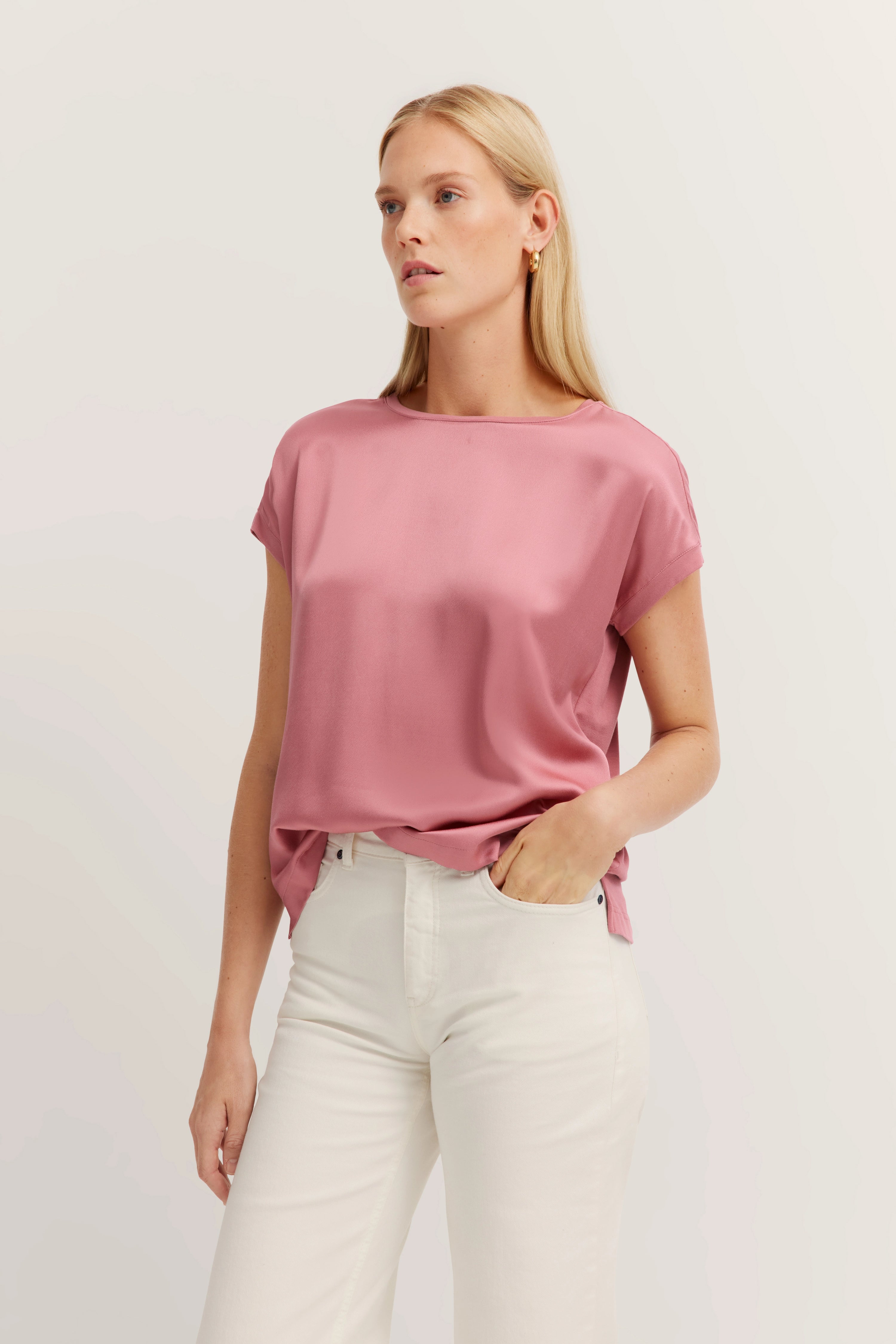 Model met roze blouse en witte broek, ontspannen pose met hand in zak.