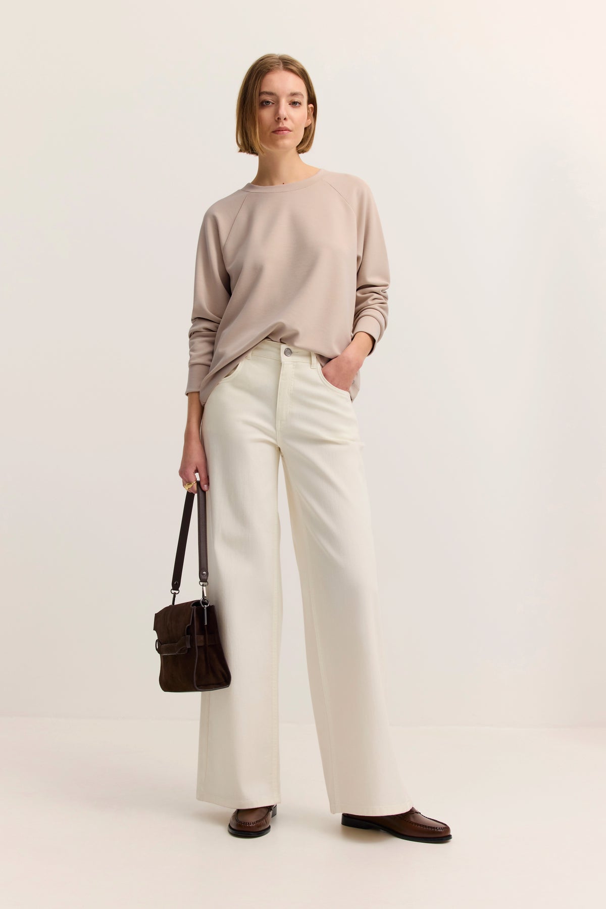 Beiger Pullover, weiße Hose, braune Tasche und Schuhe, Model stehend mit Hand in Tasche.