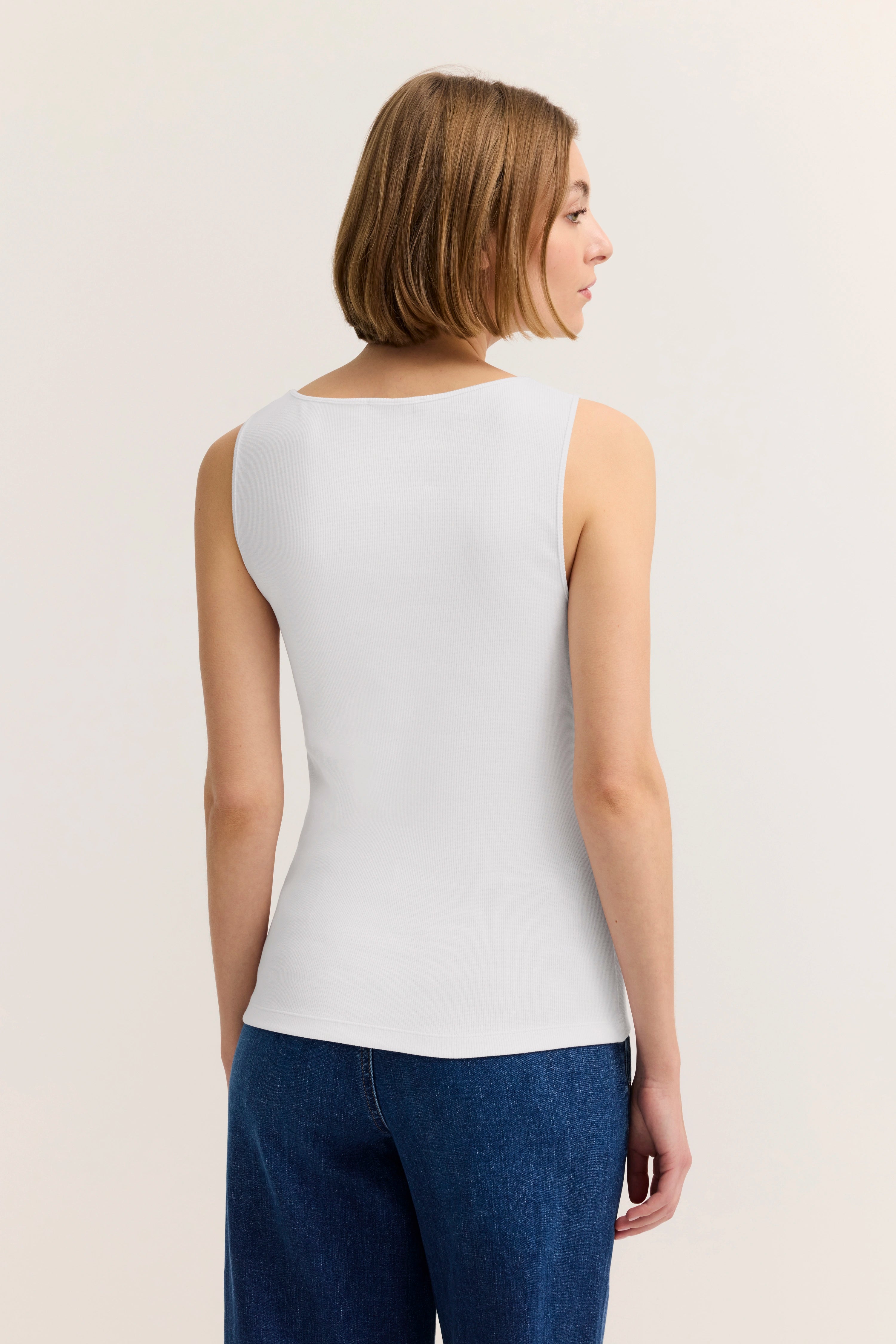 Weißes ärmelloses Top, Rückansicht, kombiniert mit blauen Jeans. Model steht seitlich.
