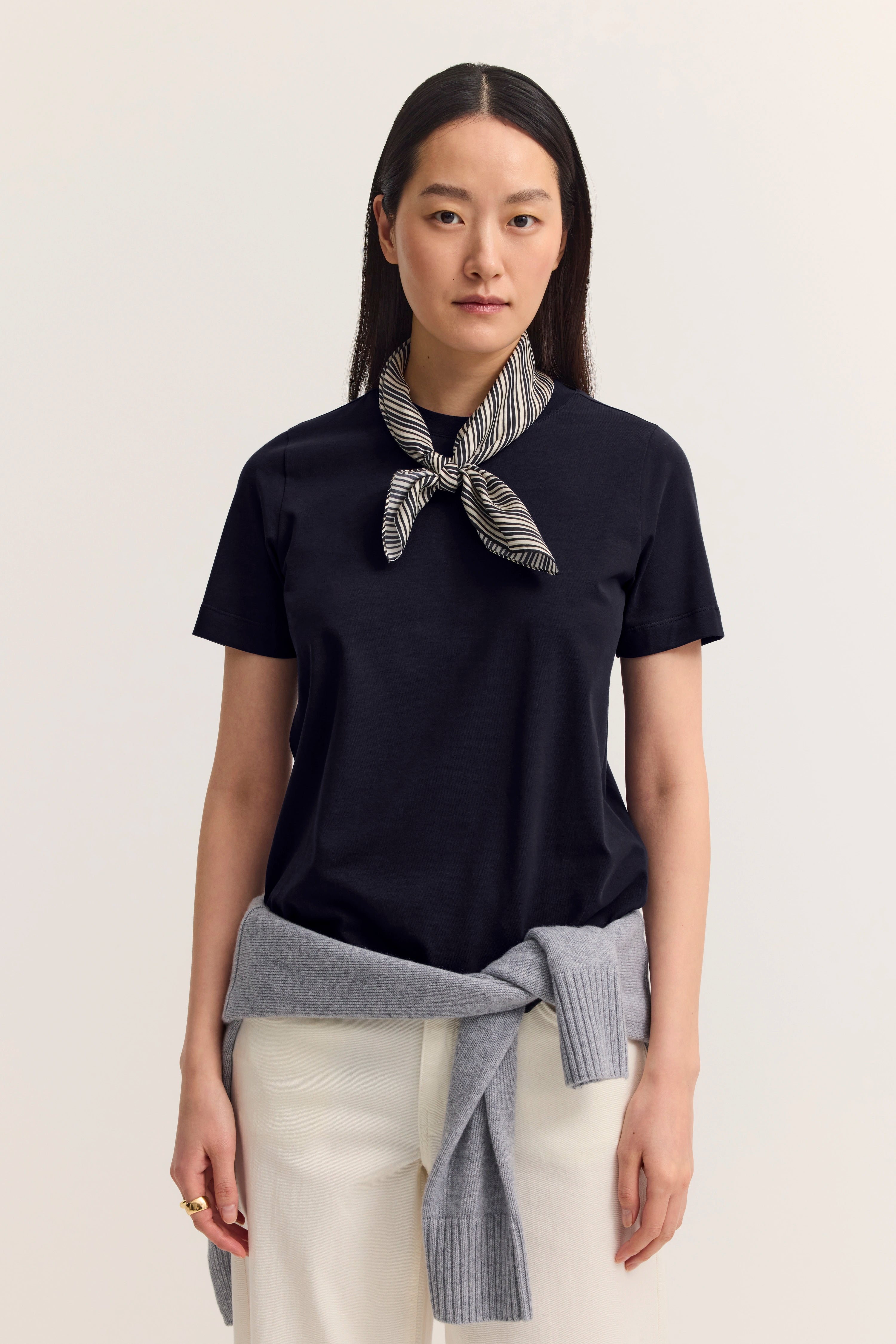 Model in navy T-Shirt mit gestreiftem Halstuch, grauer Pullover um die Taille, cremefarbene Hose.