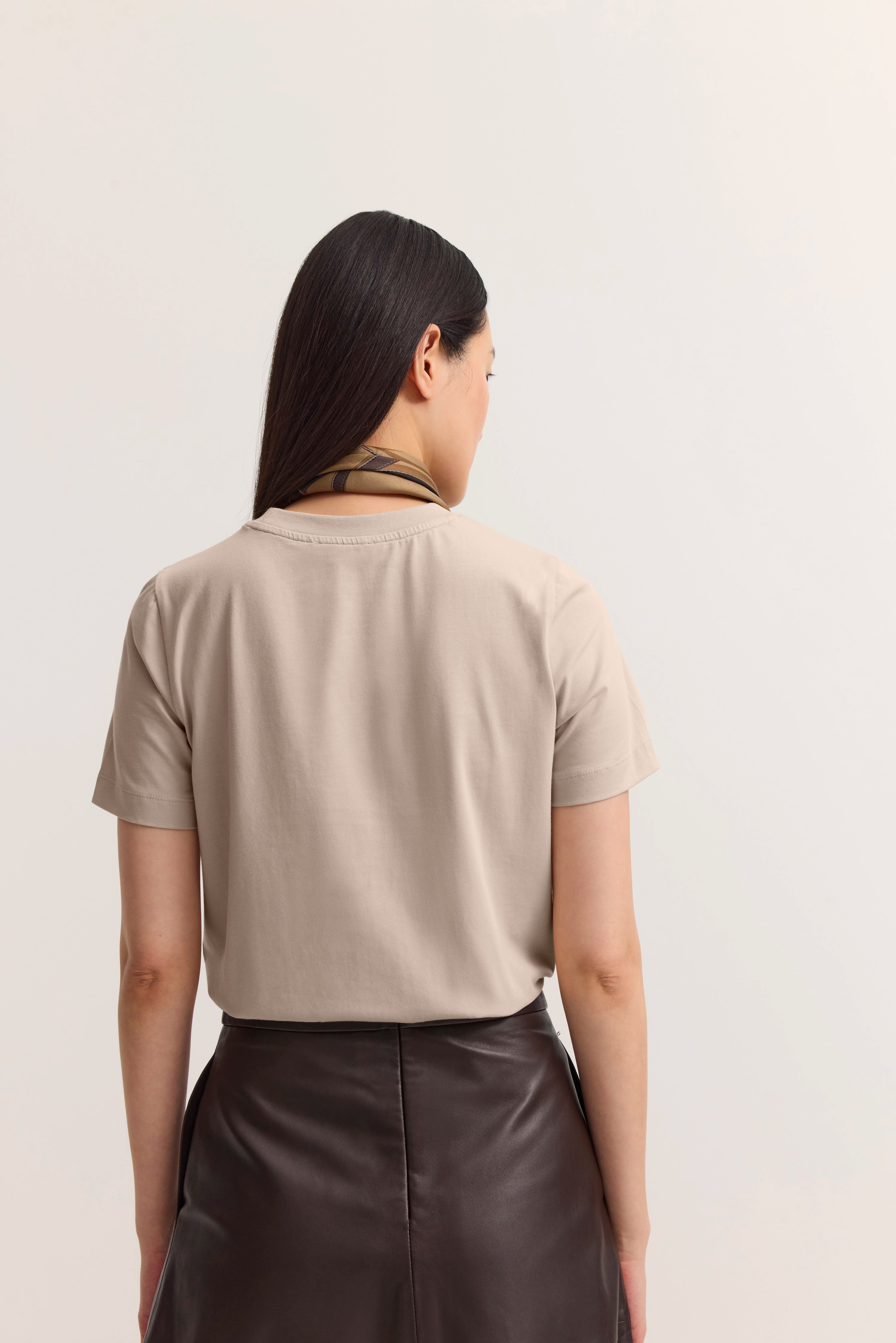 Beige T-Shirt mit braunem Lederrock, Model von hinten, entspanntes Posing.