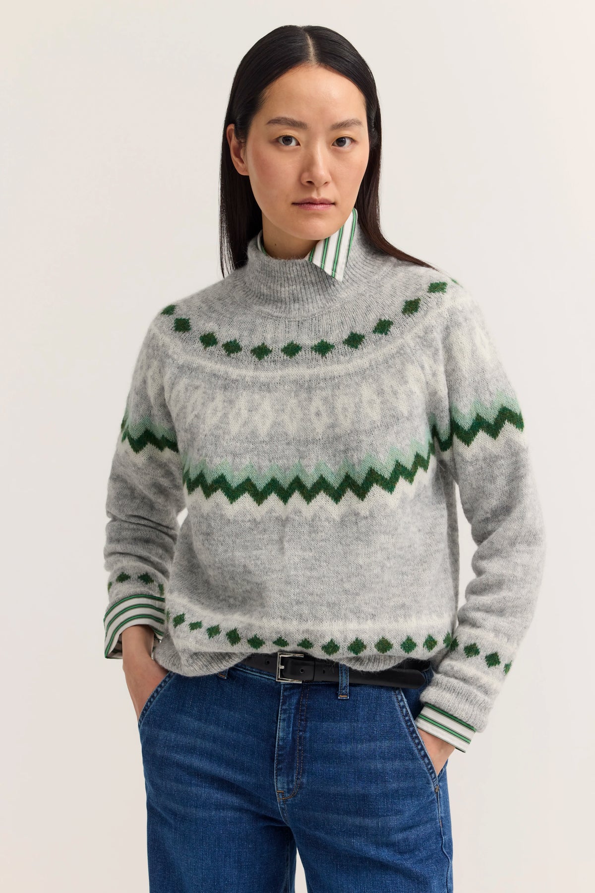 Grauer Pullover mit grünem Muster über gestreiftem Hemd, blaue Jeans, Model blickt direkt zur Kamera.