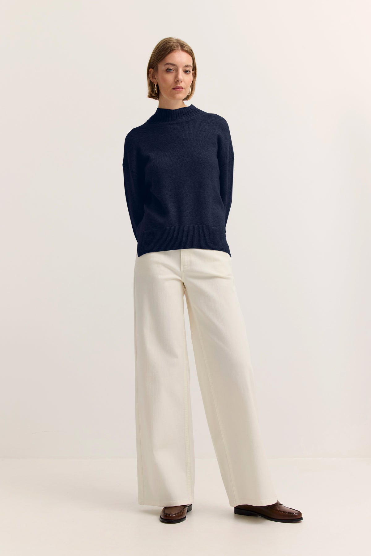 Model in dunkelblauem Pullover und weißen, weiten Hosen, steht entspannt, bugatti Stil.