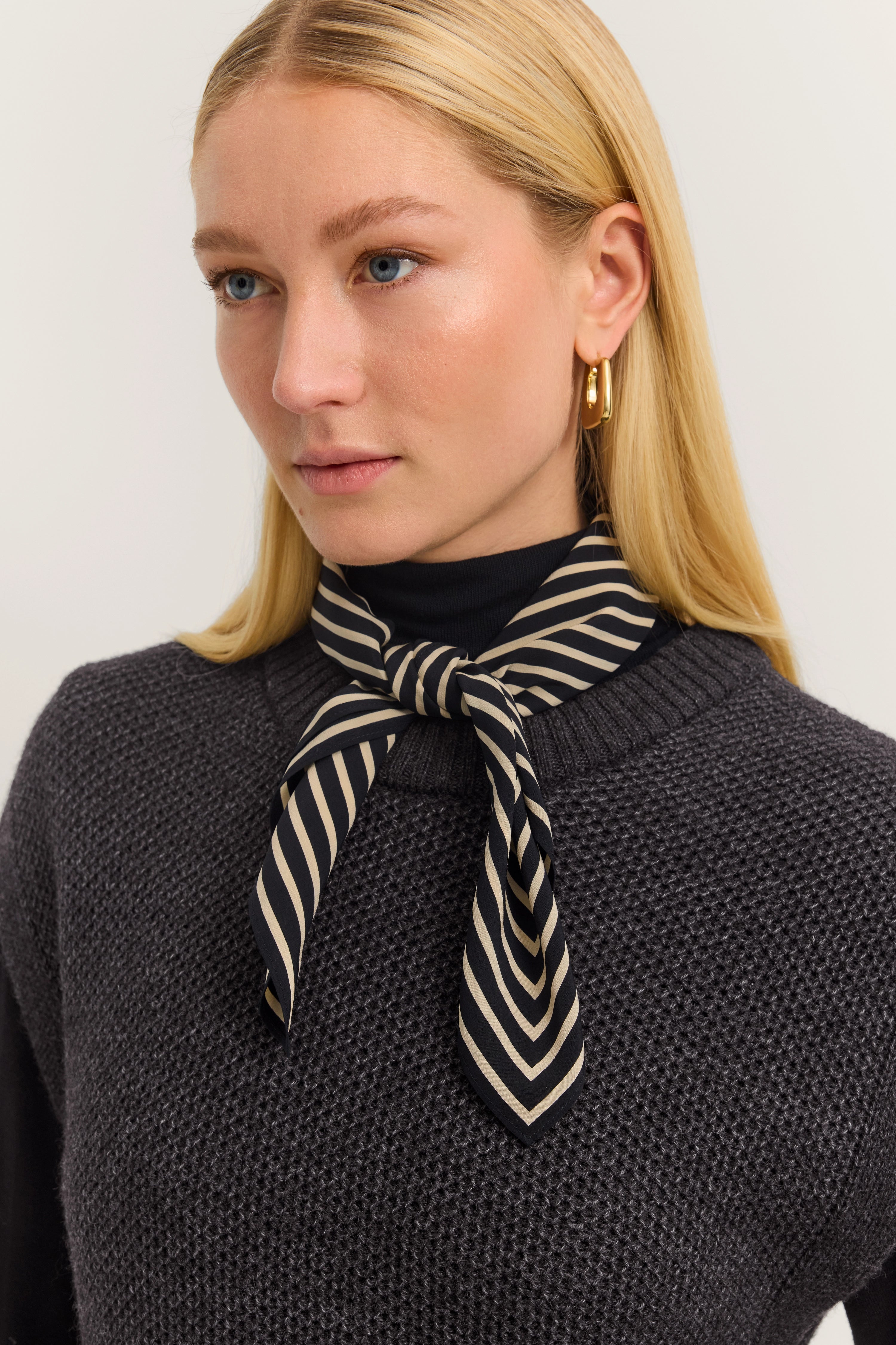Modelbild mit blondem Haar, trägt gestreiften Schal in Schwarz und Beige, dunkelgrauer Strickpullover.