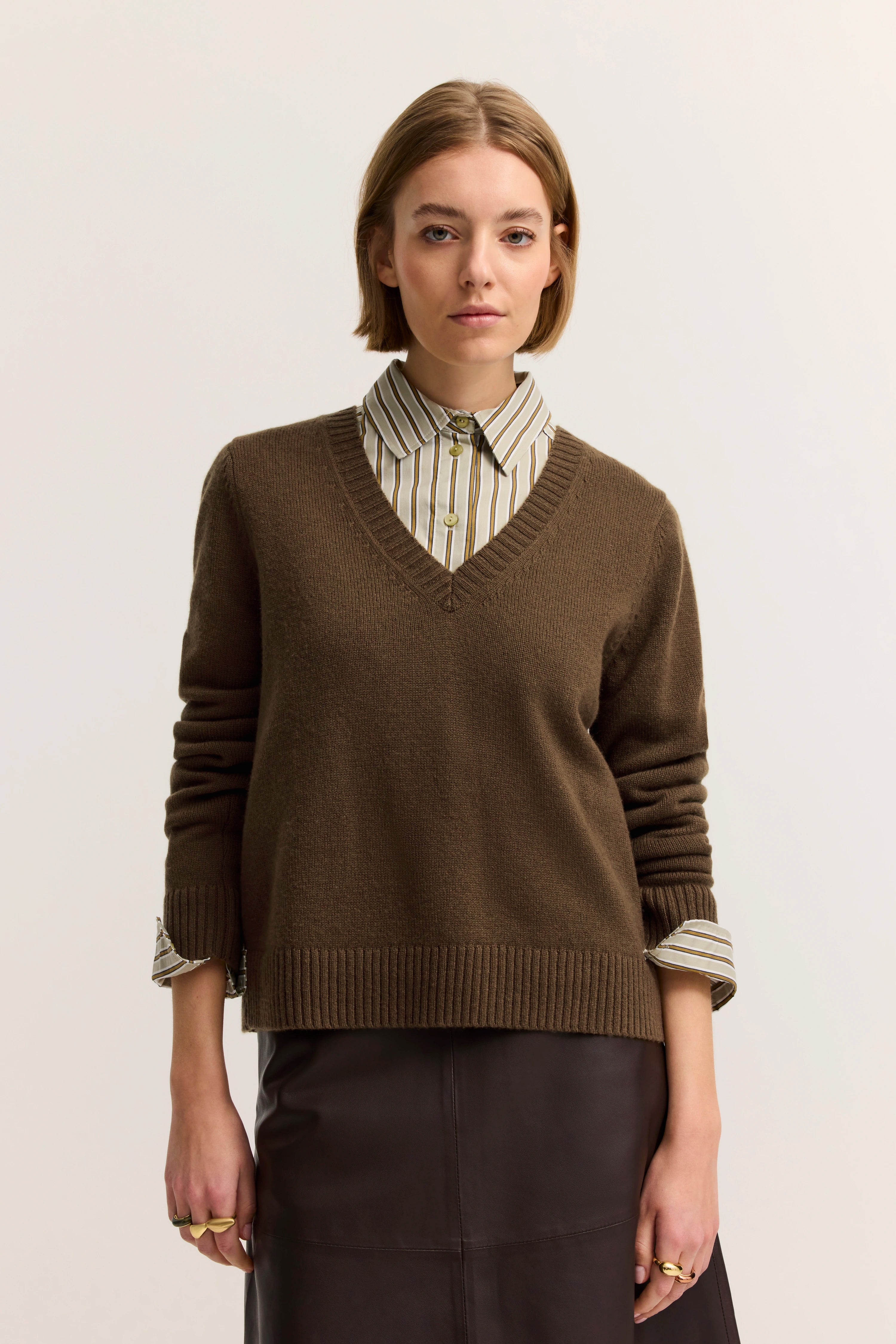 Brauner Pullover über gestreifter Bluse, Modelbild, neutraler Gesichtsausdruck, entspannter Stand.