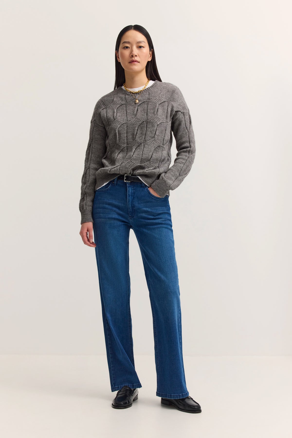 Grauer Zopfpullover und blaue Jeans, Model stehend, Hände in den Taschen.