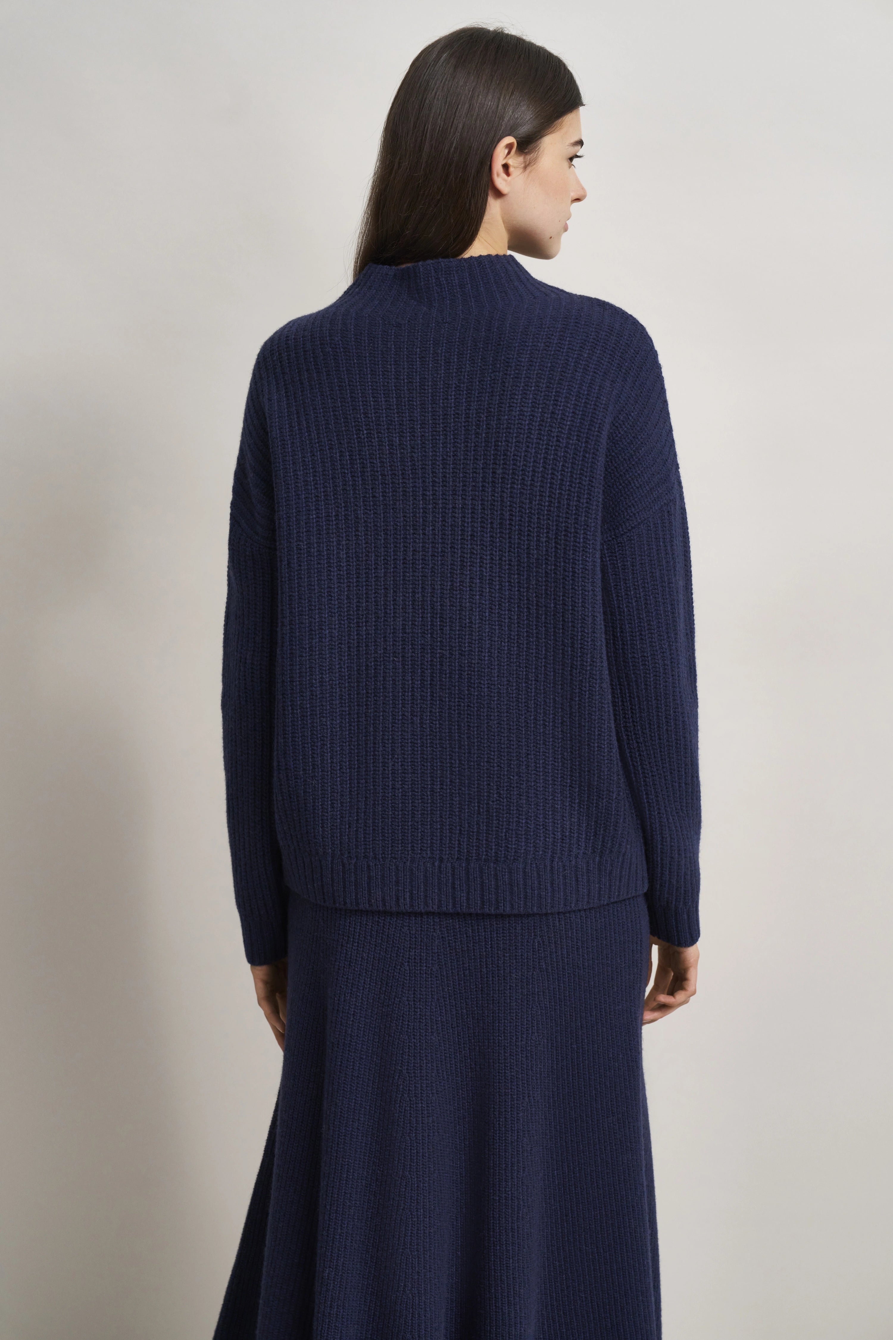 Model in dunkelblauem Strick-Pullover und Rock von hinten, legeres Standbild.