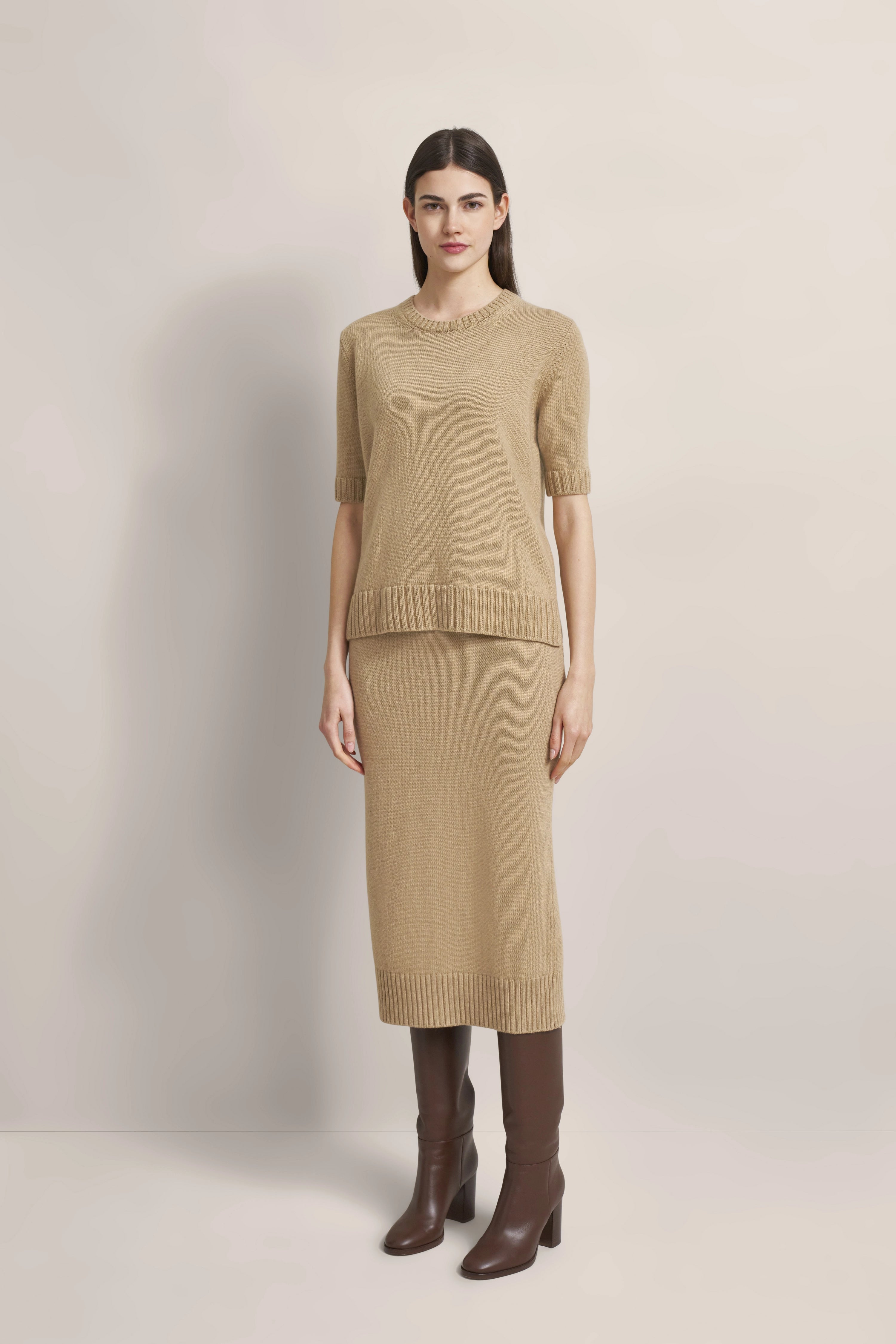 Model trägt beige Strickkleid mit kurzen Ärmeln, kombiniert mit braunen Stiefeln. Stehende Pose.