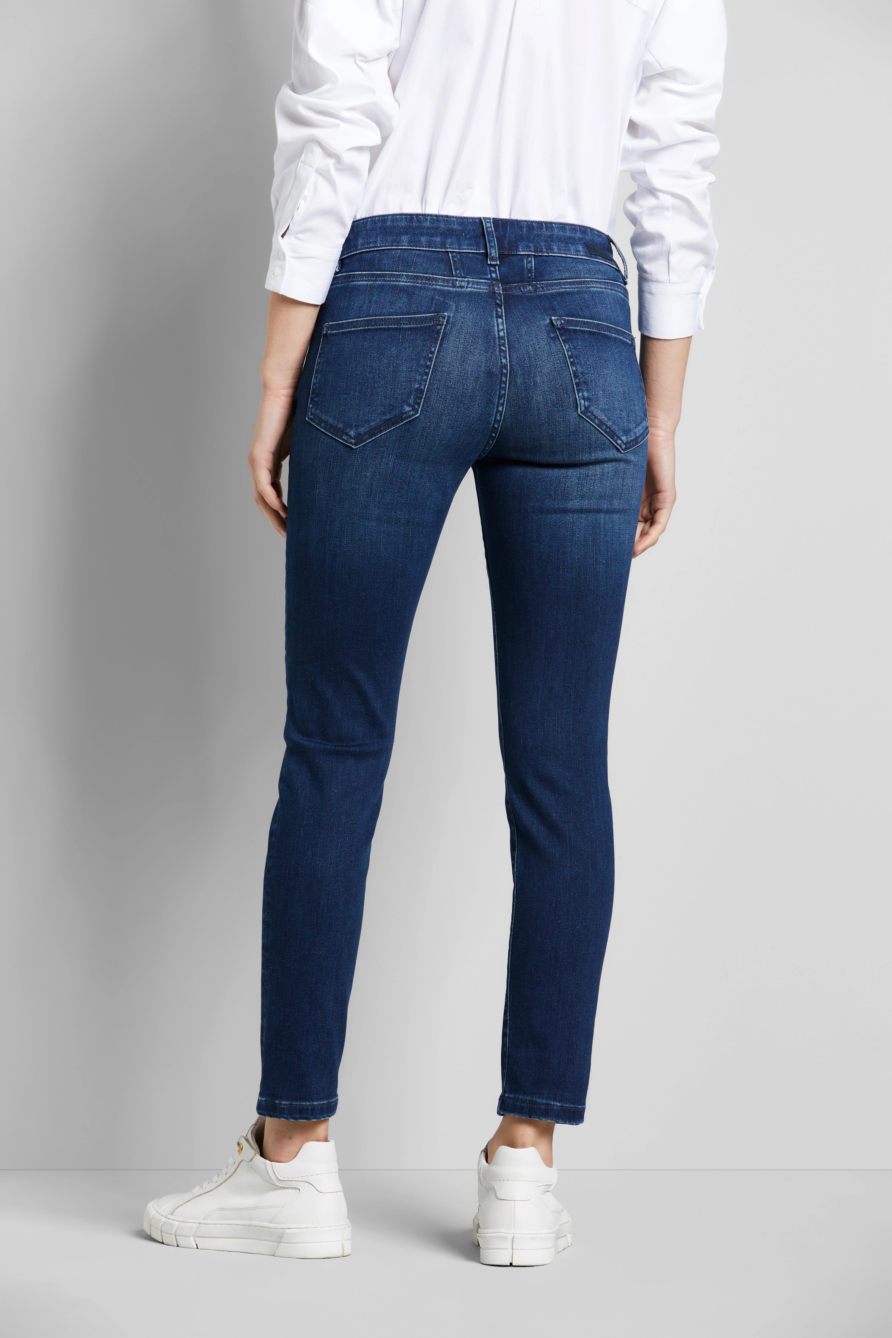 Rückansicht: Model trägt weiße Bluse, blaue Jeans von bugatti, weiße Sneaker. Entspanntes, legeres Outfit.